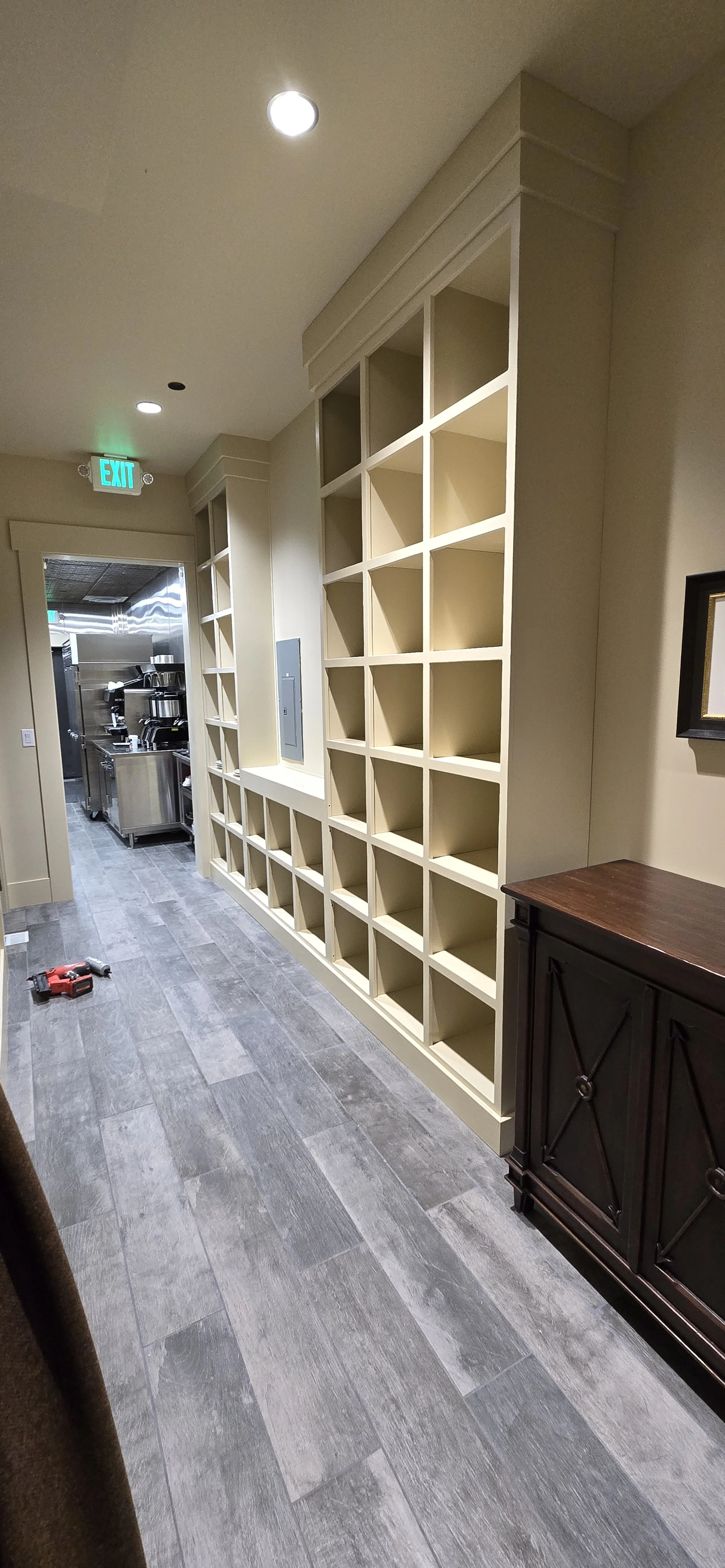 Empty beige shelving unit with multiple cubbies in a corridor, with a dark brown cabinet to the right and a kitchenette with appliances in the background.