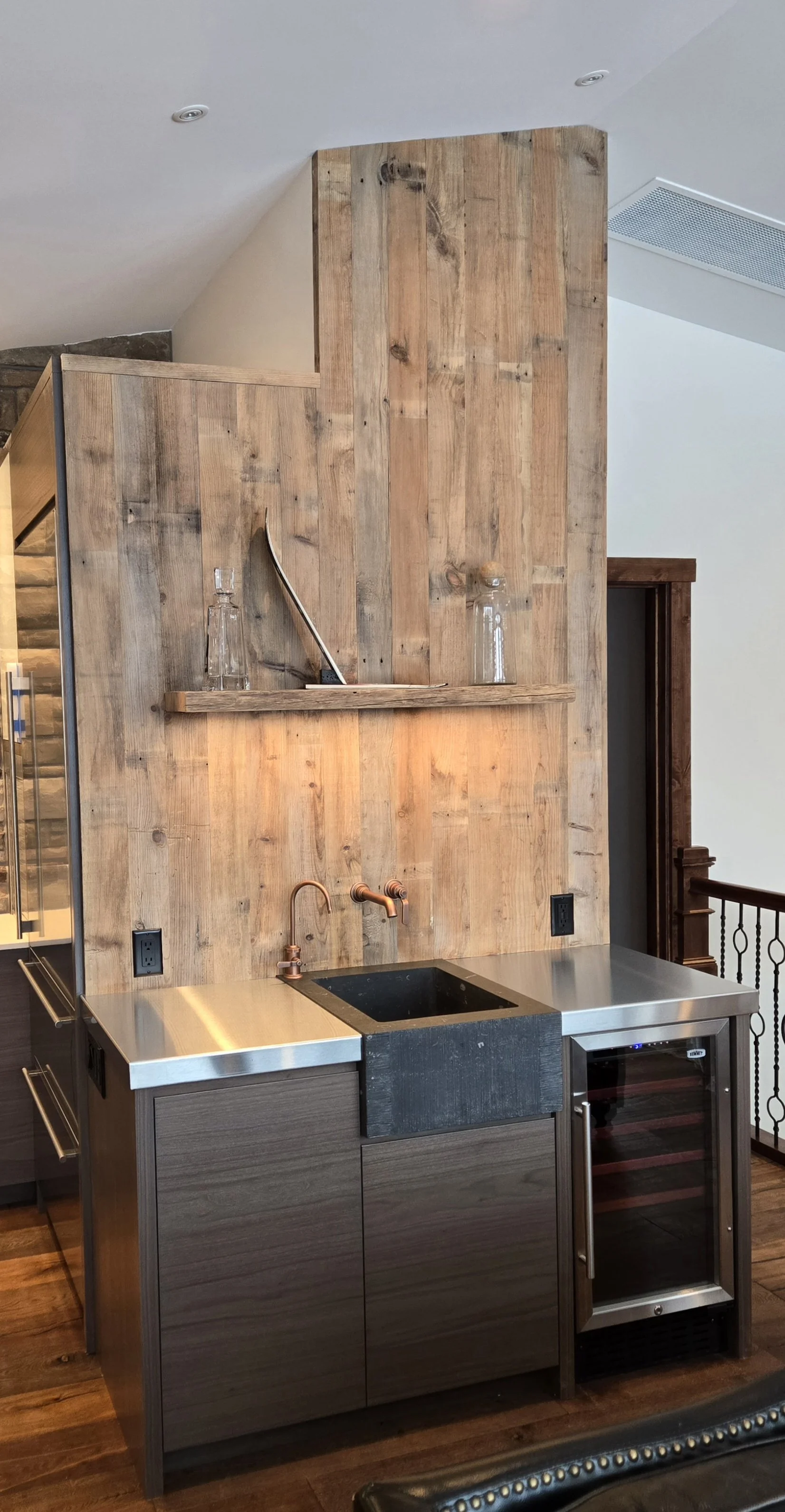 A modern bar area with a wooden panel wall, copper faucet, black sink, stainless steel countertop, and a small wine fridge.