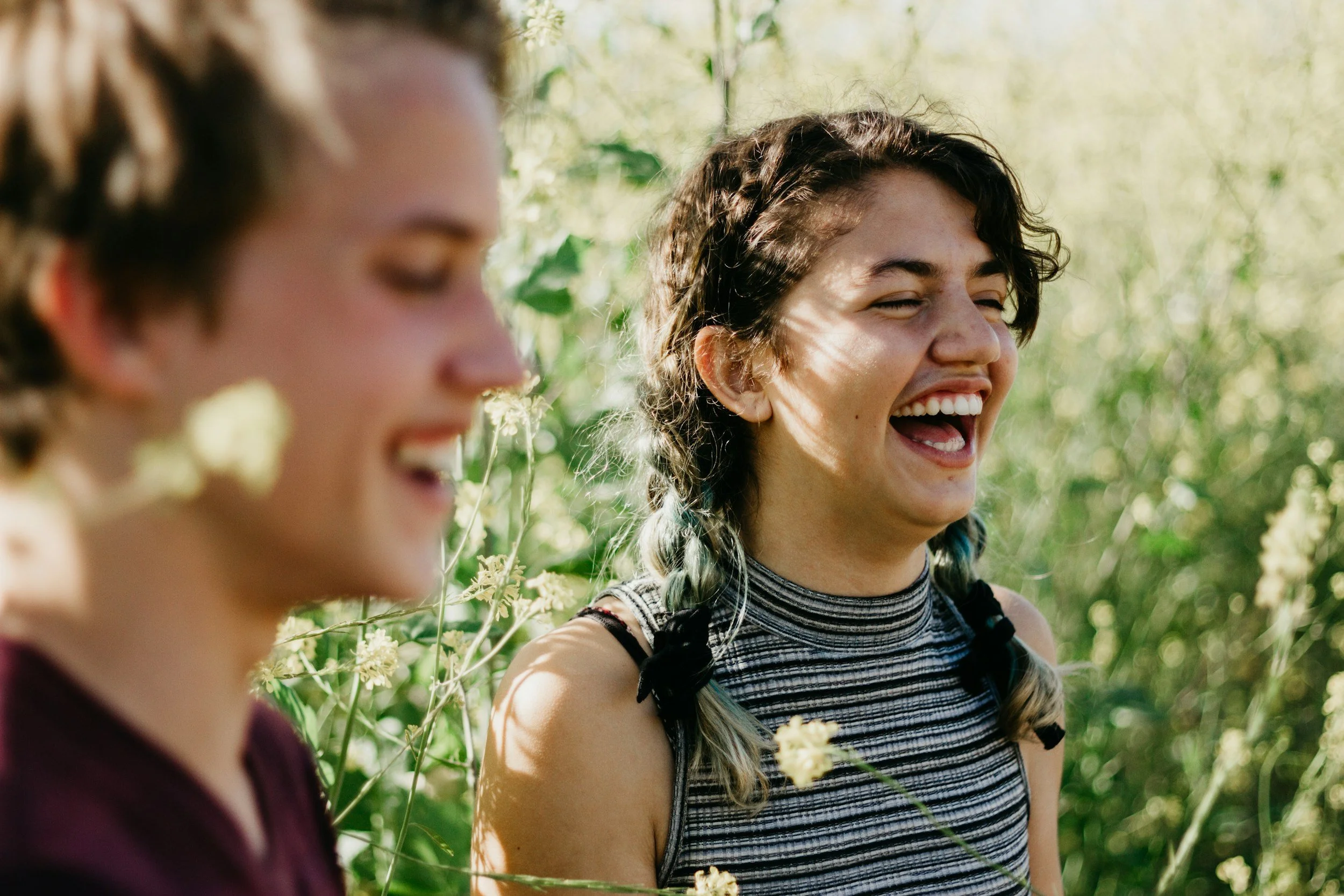 Two young women laughing outdoors among green plants and flowers, sunlight filtering through the foliage.