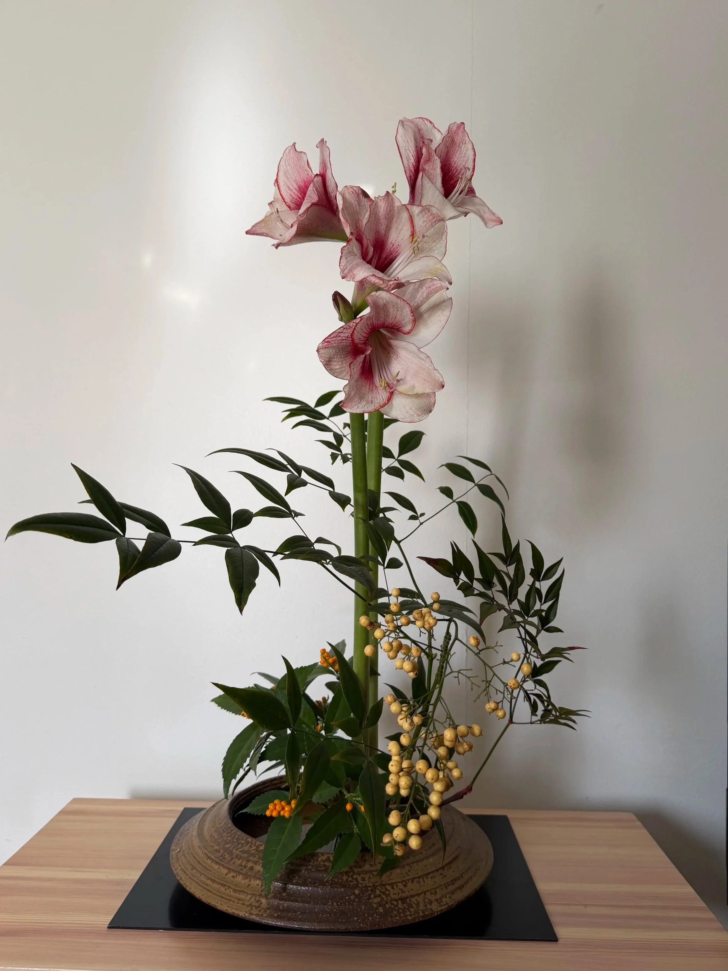 A floral arrangement with tall pink and white amaryllis flowers, green leaves, and yellow berries in a round, textured brown vase on a wooden table against a plain white wall.