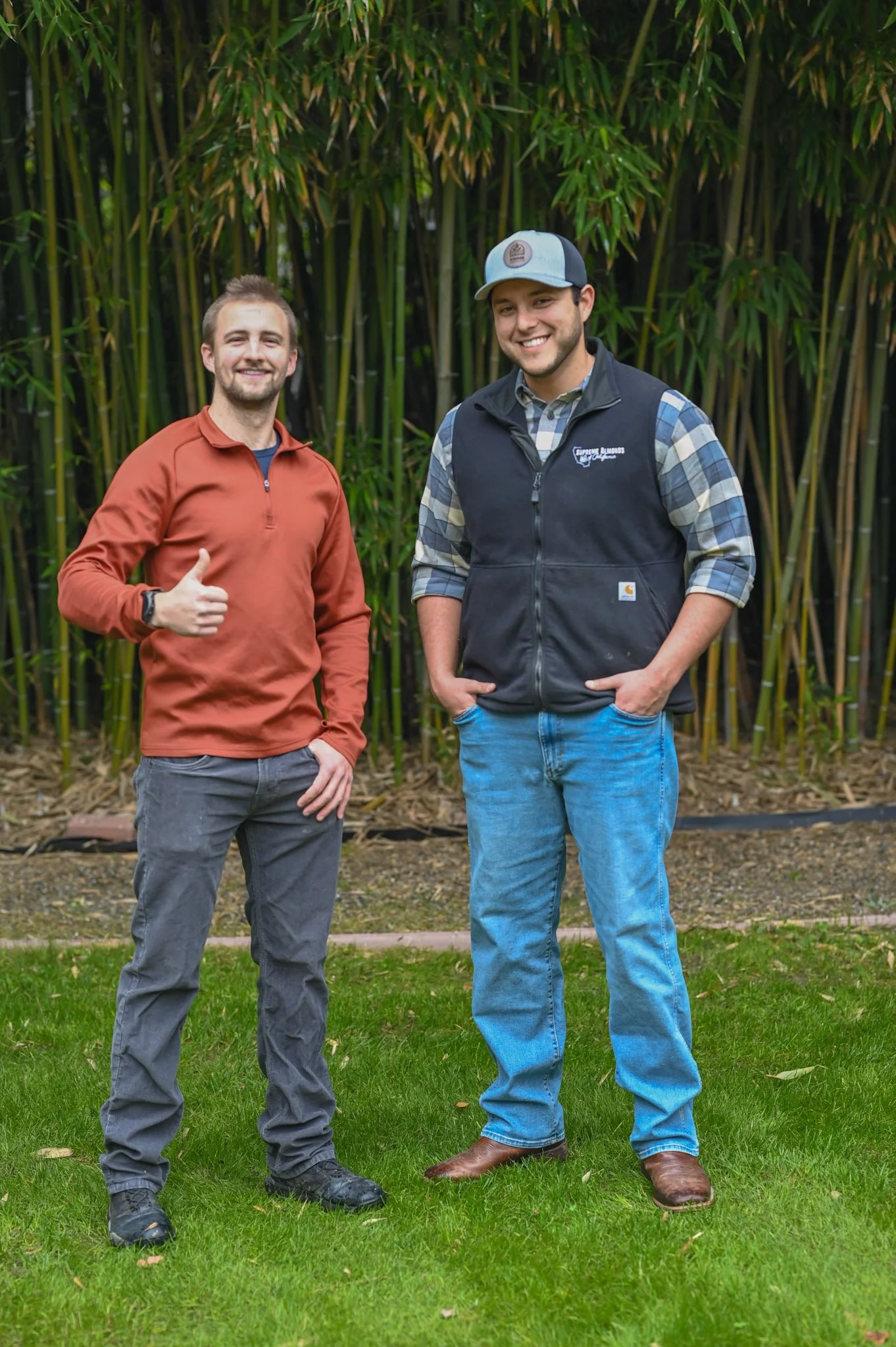 Two men standing outdoors in front of tall green bamboo, smiling at the camera. The man on the left is wearing a reddish-brown zip-up jacket, gray pants, and black shoes, giving a thumbs-up. The man on the right is wearing a plaid shirt, a black vest, blue jeans, brown boots, and a gray and black cap.