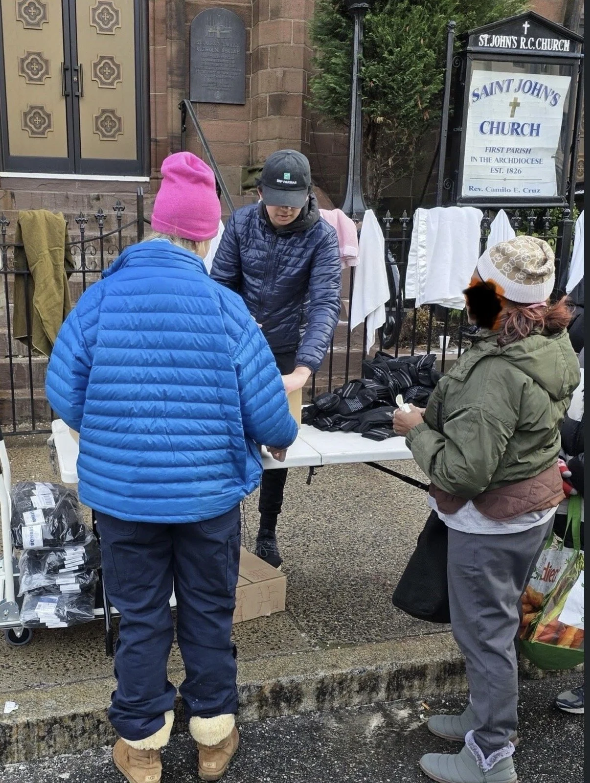 People shopping for clothing outside of Saint John's Church, with several clothing items on display and hanging towels behind them.