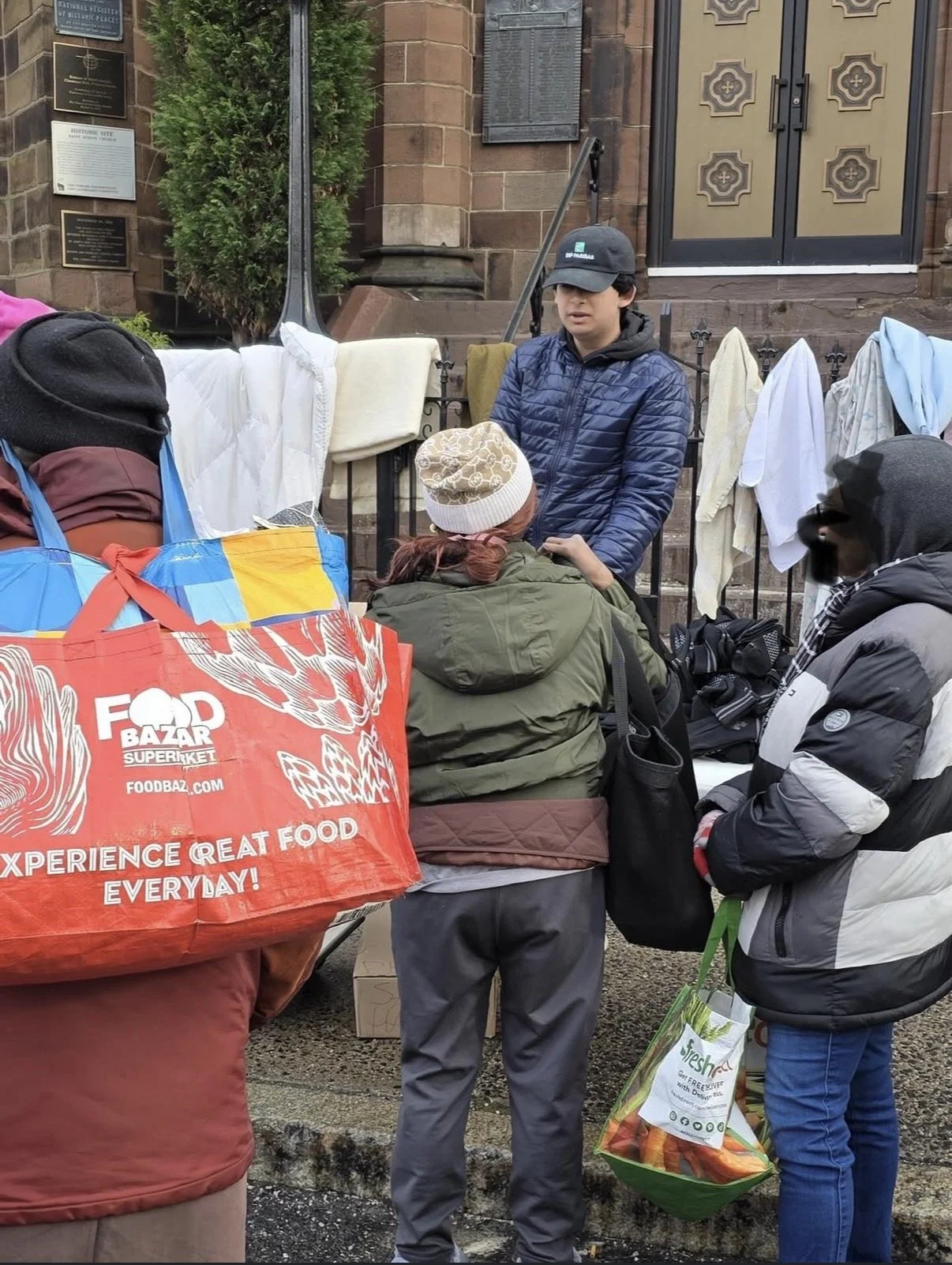 People standing in line at an outdoor market booth with a person attending, against a background of a brick building with historic plaques. The people are dressed in jackets and hats, carrying shopping bags, one of which reads 'FOOD BAZAR'.