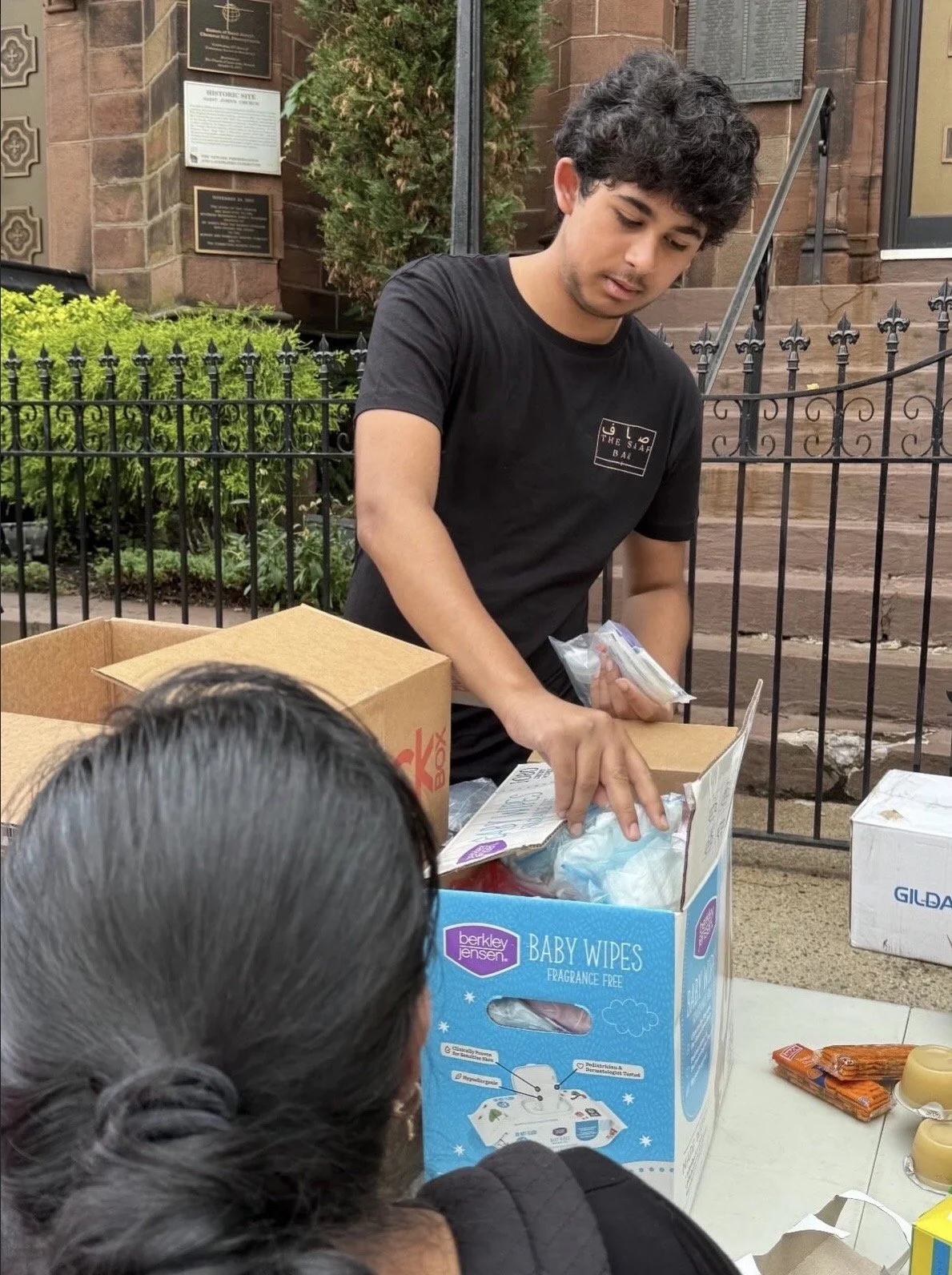 A young man with dark, curly hair wearing a black T-shirt handing out supplies from a box labeled baby wipes to a woman with long, dark hair. They are outdoors near a brick building with steps and a black fence.