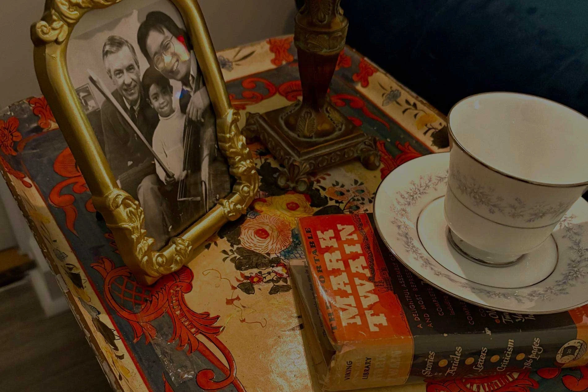 A framed black and white photograph of three people, two men and a boy, is placed on a colorful tablecloth. The table also has two stacked teacups with floral patterns and a table lamp with an ornate base. A vintage book is partially visible beneath the tablecloth.