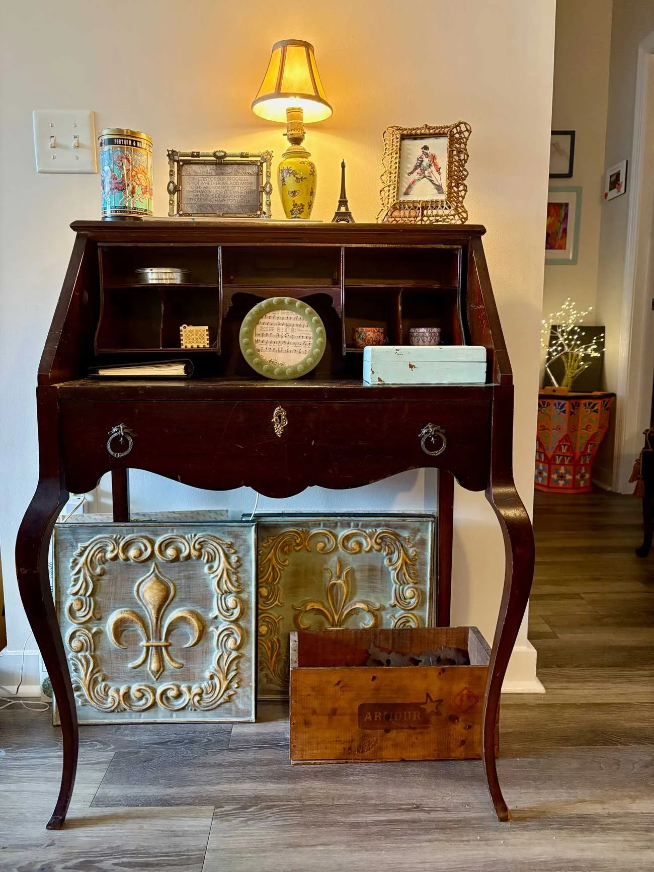 A vintage wooden Secretary desk with various decorative items on top, including a lamp, photo frames, a miniature Eiffel Tower, and other small objects. The desk has ornate drawer fronts with a fleur-de-lis design, and there is a decorative metal box on the floor in front of it. To the right, a colorful patterned basket is visible, and the background includes a hallway with framed artwork on the walls and a lit decorative tree.