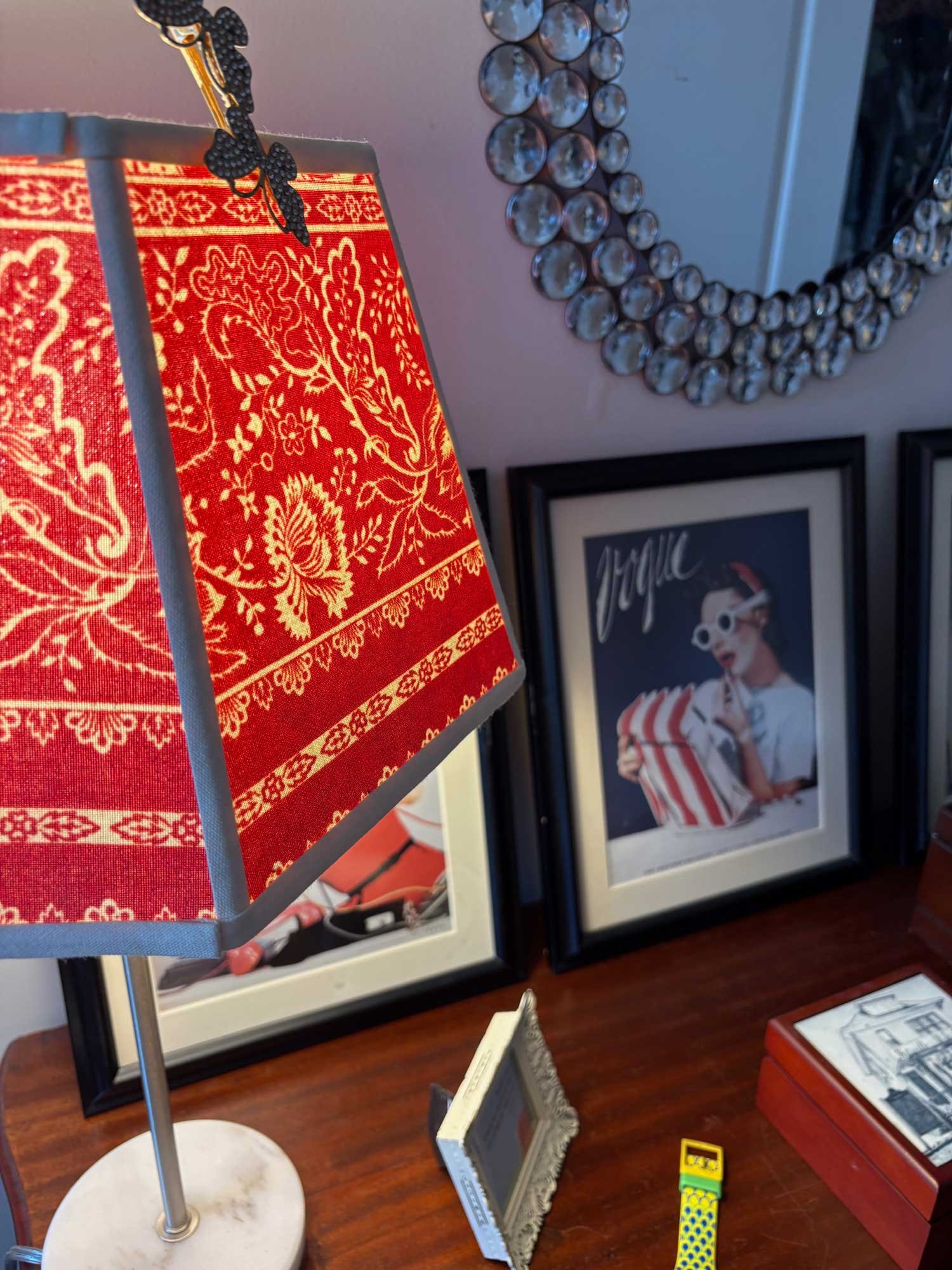 Deco lamp with red patterned shade, round marble base, and metallic frog decoration on top, with framed art and small toys on a wooden surface in the background.