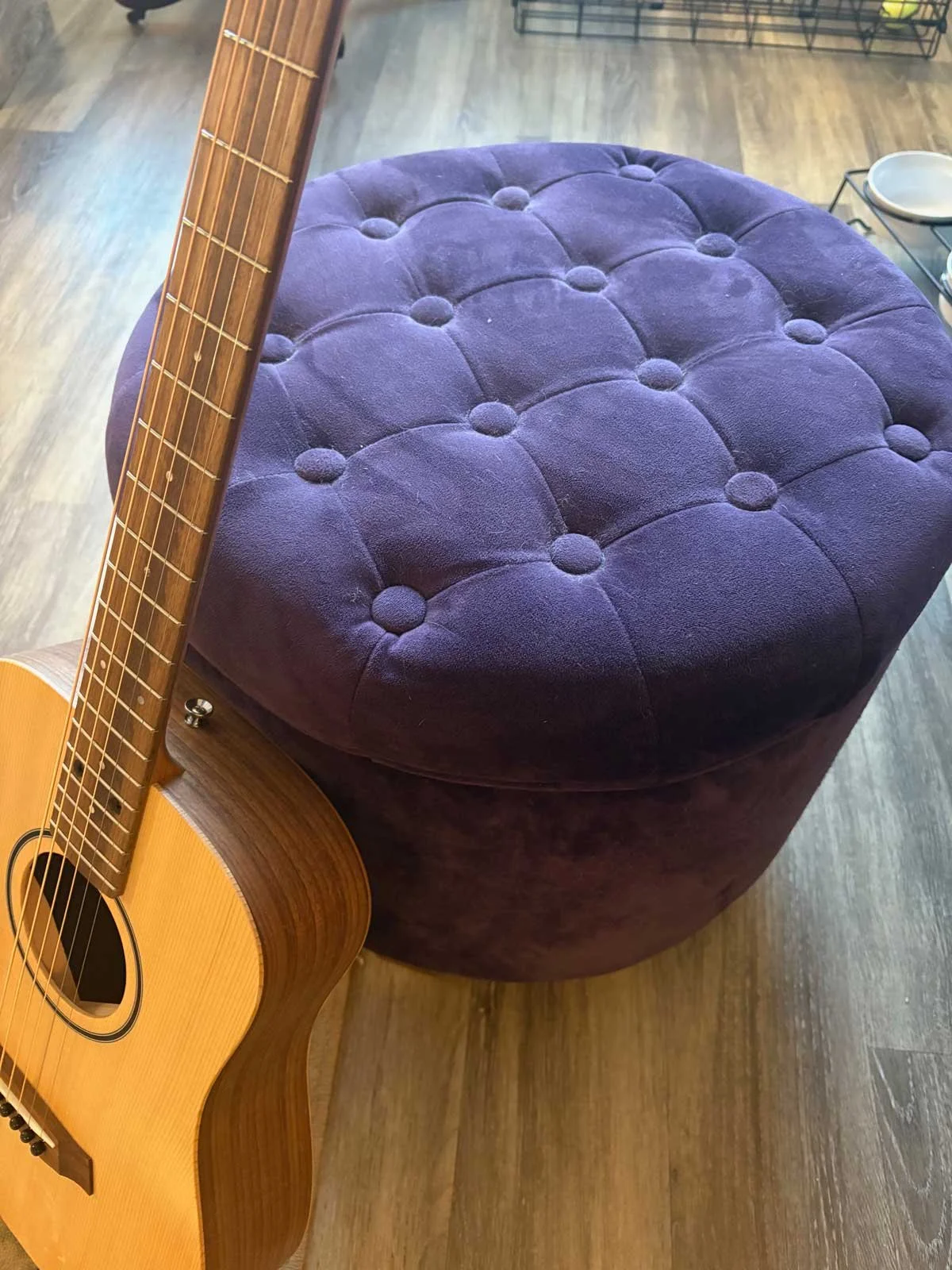 An acoustic guitar leaning against a round, purple, tufted ottoman on a wooden floor.
