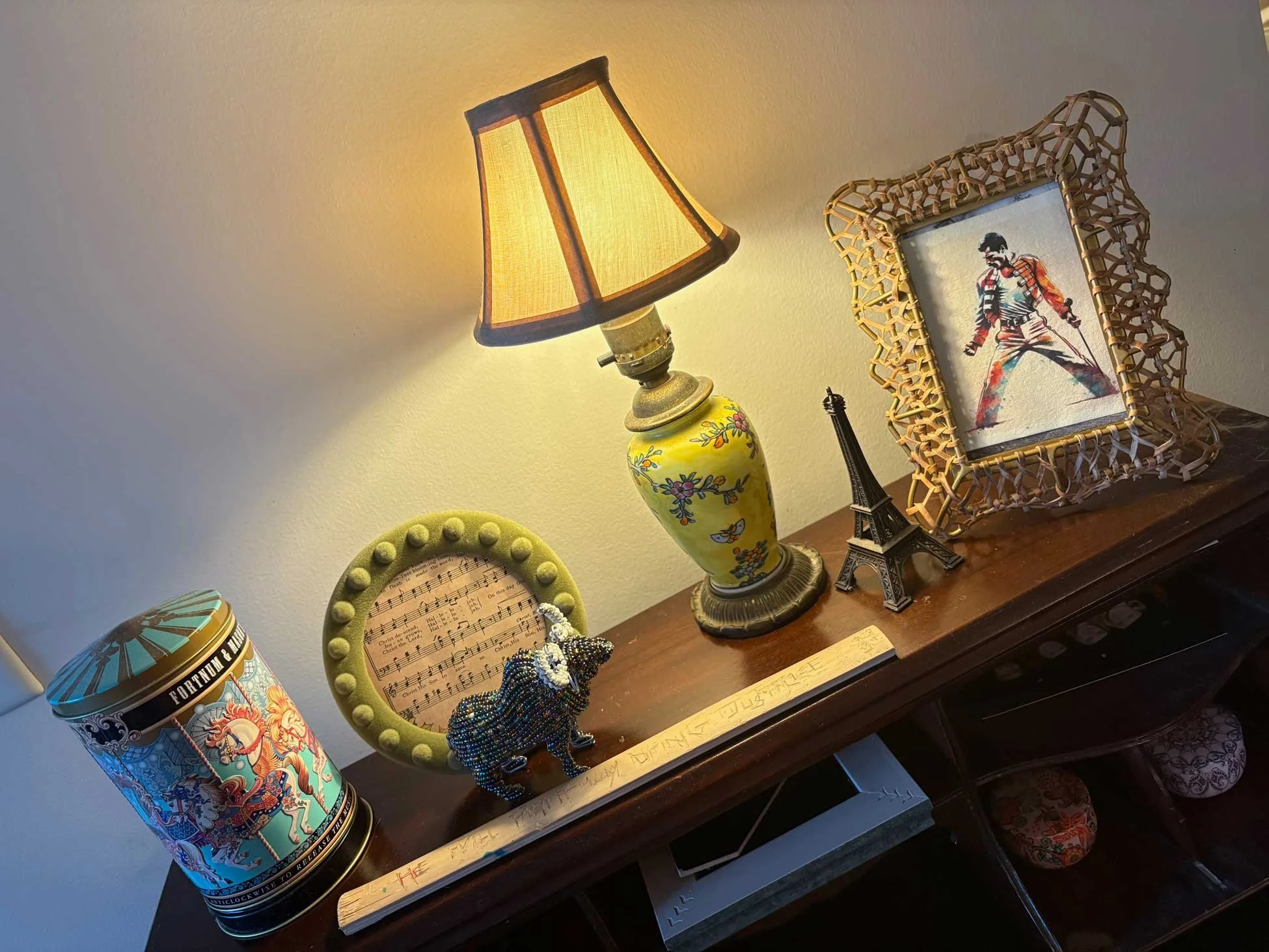 A wooden table against a white wall displays various decorative items: a vintage-style yellow table lamp with a beige fabric shade, a framed picture of a martial artist, a small model of the Eiffel Tower, a music page in a green frame, a beaded elephant figurine, a round container with a colorful design, and a few other small items and notes.