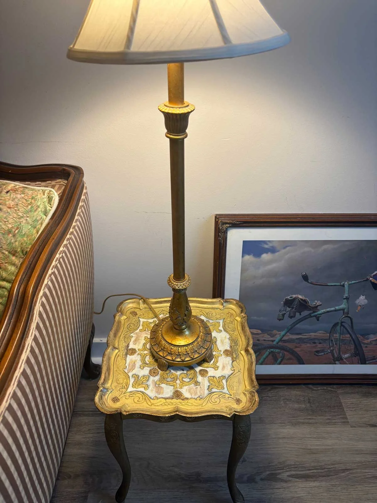 An ornate side table with a gold and cream design, holding a vintage floor lamp with a beige lampshade. To the right, a framed photograph of a bicycle outdoors. Part of a striped upholstered sofa with a floral pillow is visible on the left.