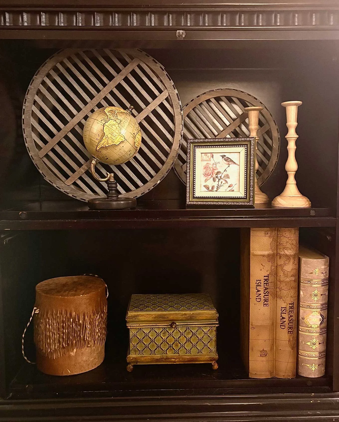 Decorative shelves with a globe, framed bird and floral artwork, wooden candlesticks, antique books, a small box, and a textured brown jar.