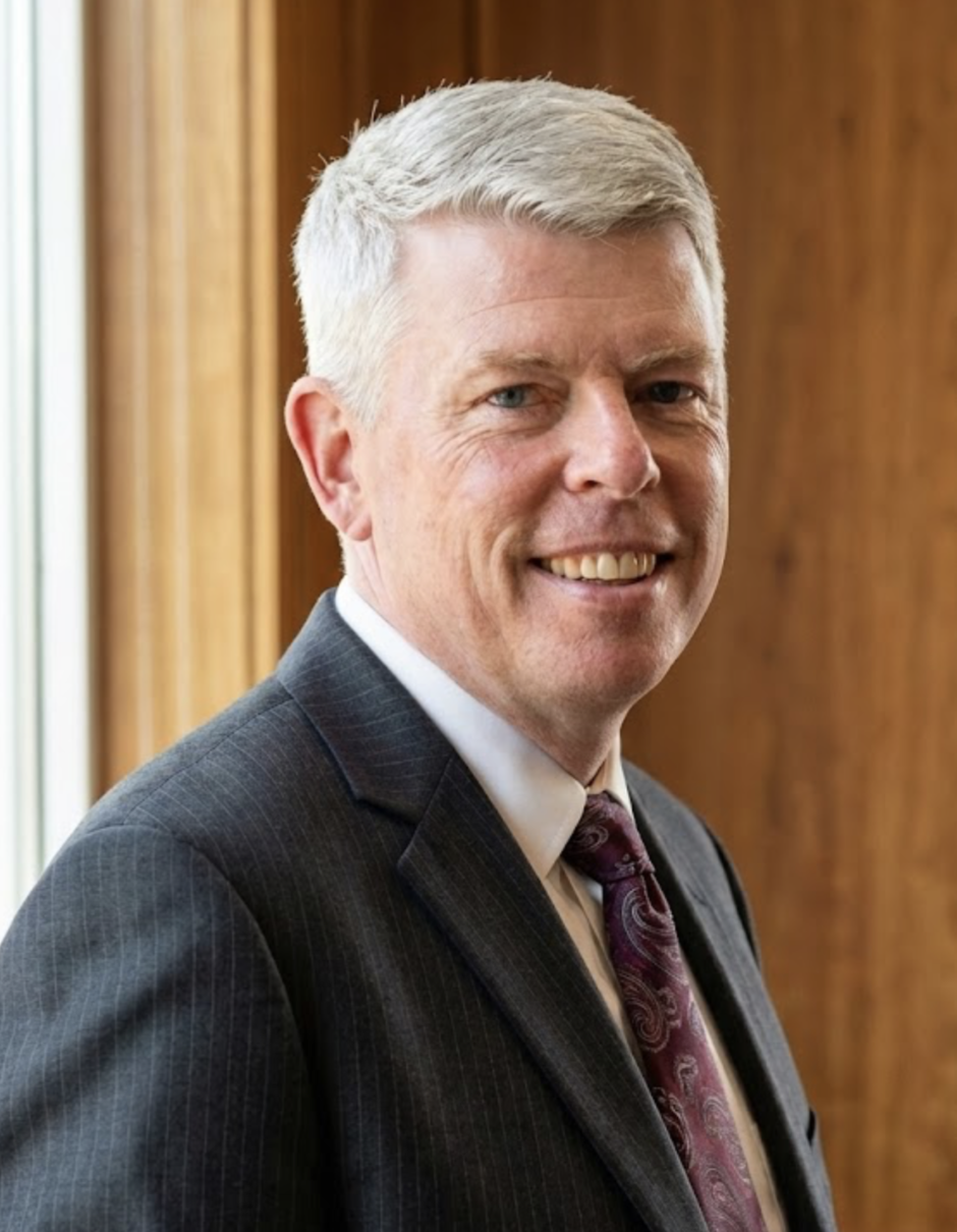 A middle-aged man with short gray hair in a business suit, smiling, standing indoors with wooden wall background.