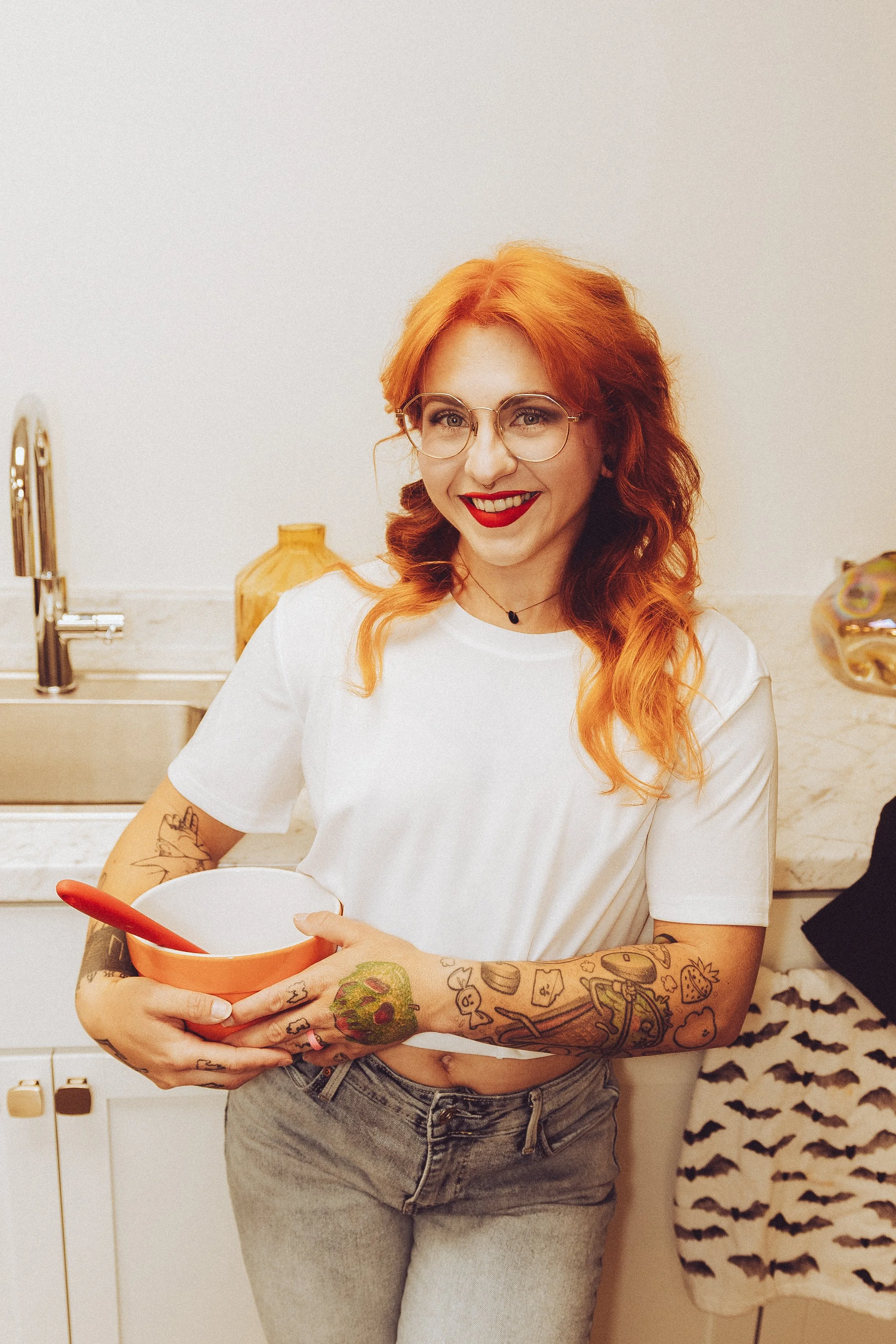 A woman with red hair, glasses, and tattoos on her arms, wearing a white t-shirt and gray jeans, standing in a kitchen and smiling while holding a bowl and spoon.