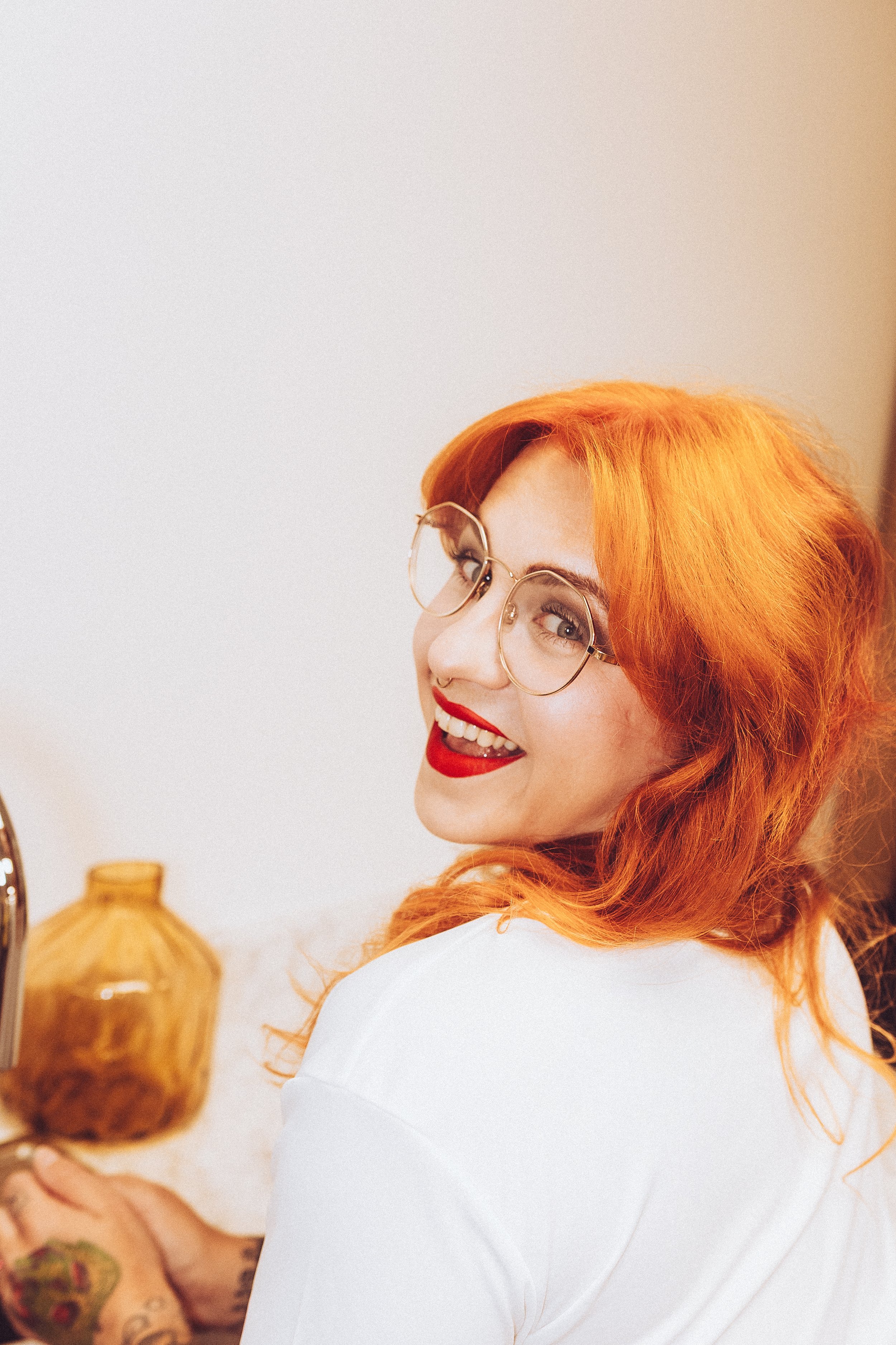 A woman with vibrant red hair, glasses, and red lipstick is smiling at the camera in an indoor setting, with a blurred background featuring a decorative vase.