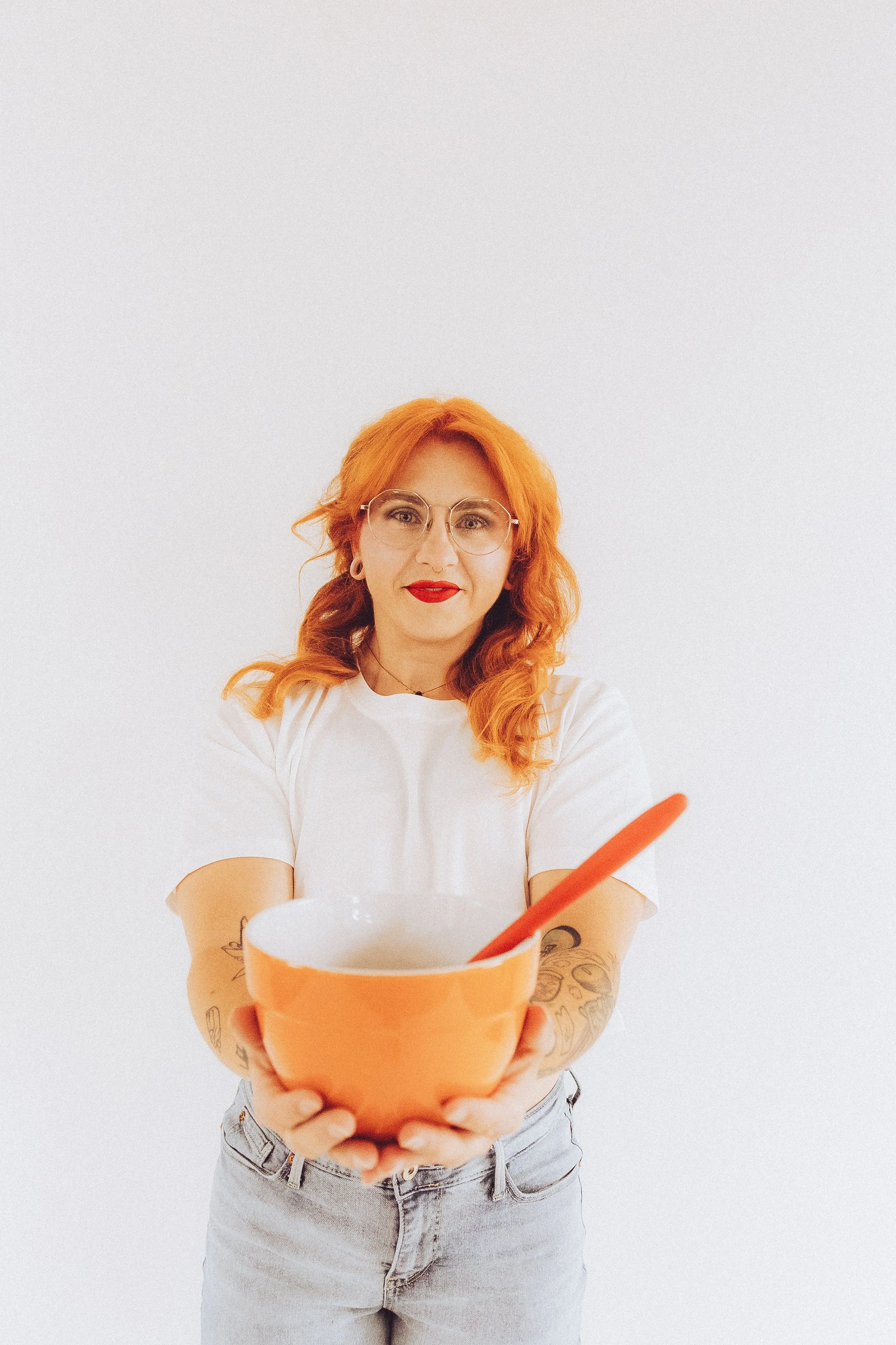 A woman with red hair, glasses, and tattoos wearing a white t-shirt and jeans, holding an orange bowl with a spoon, standing against a white background.