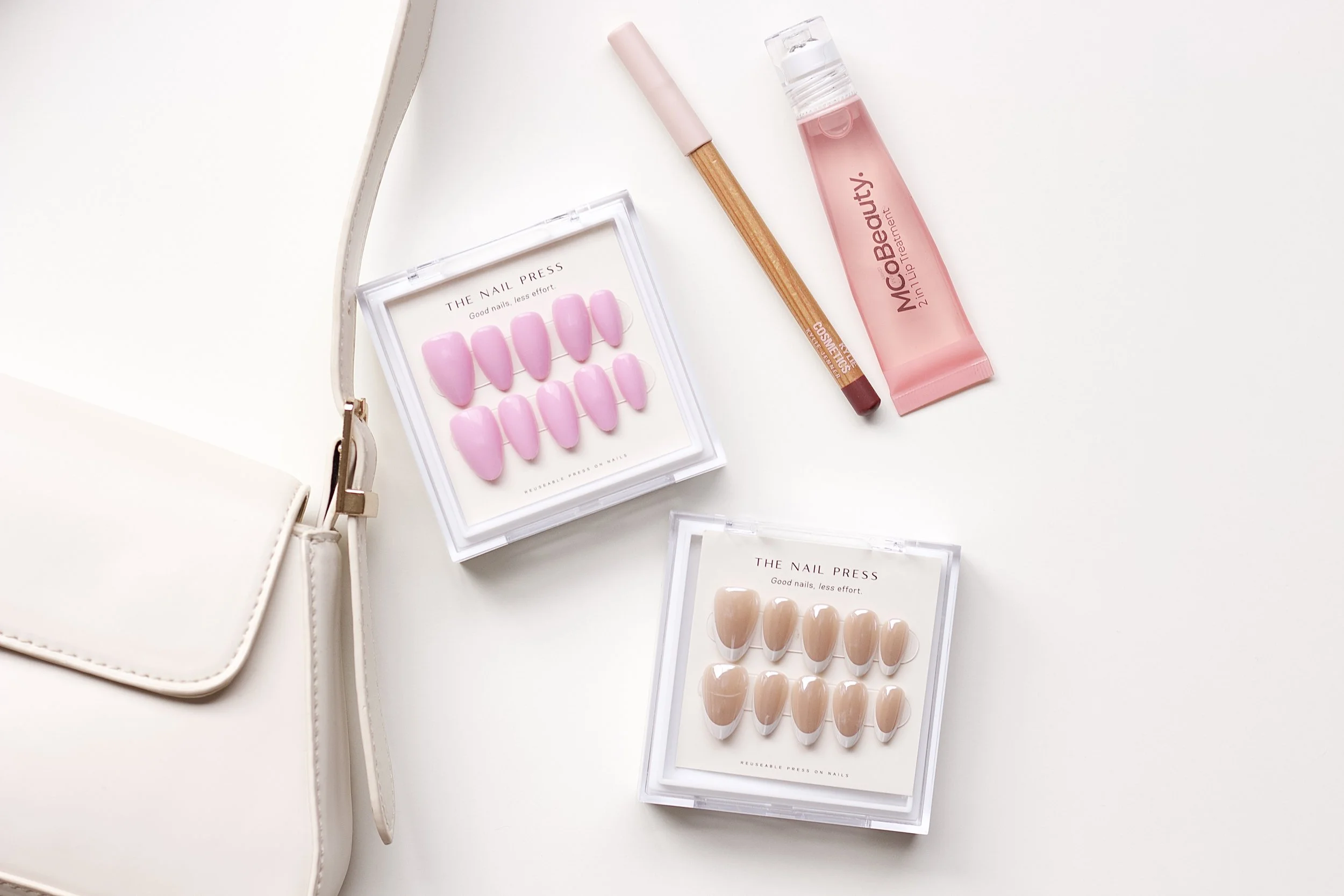A white surface with a white purse, nail press sets in pink and beige, a pink lip cream, and a pink lip gloss in a clear bottle.