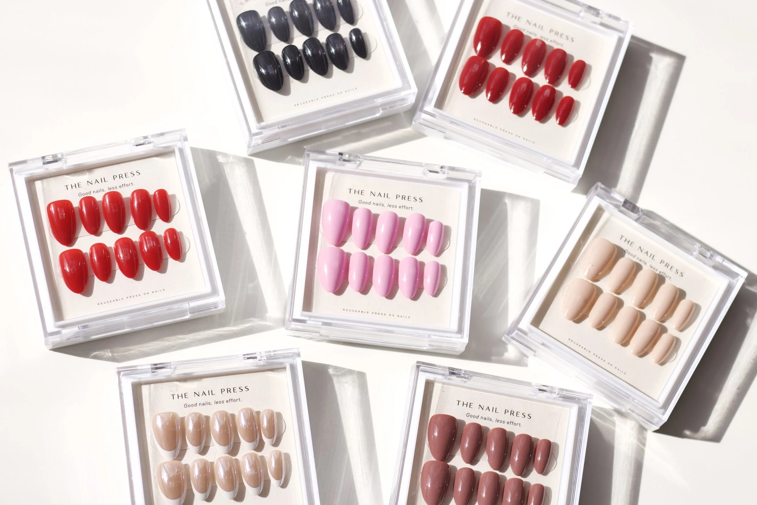 Multiple packs of fake nails in various colors including black, red, pink, beige, and mauve, displayed in clear plastic cases on a white surface.