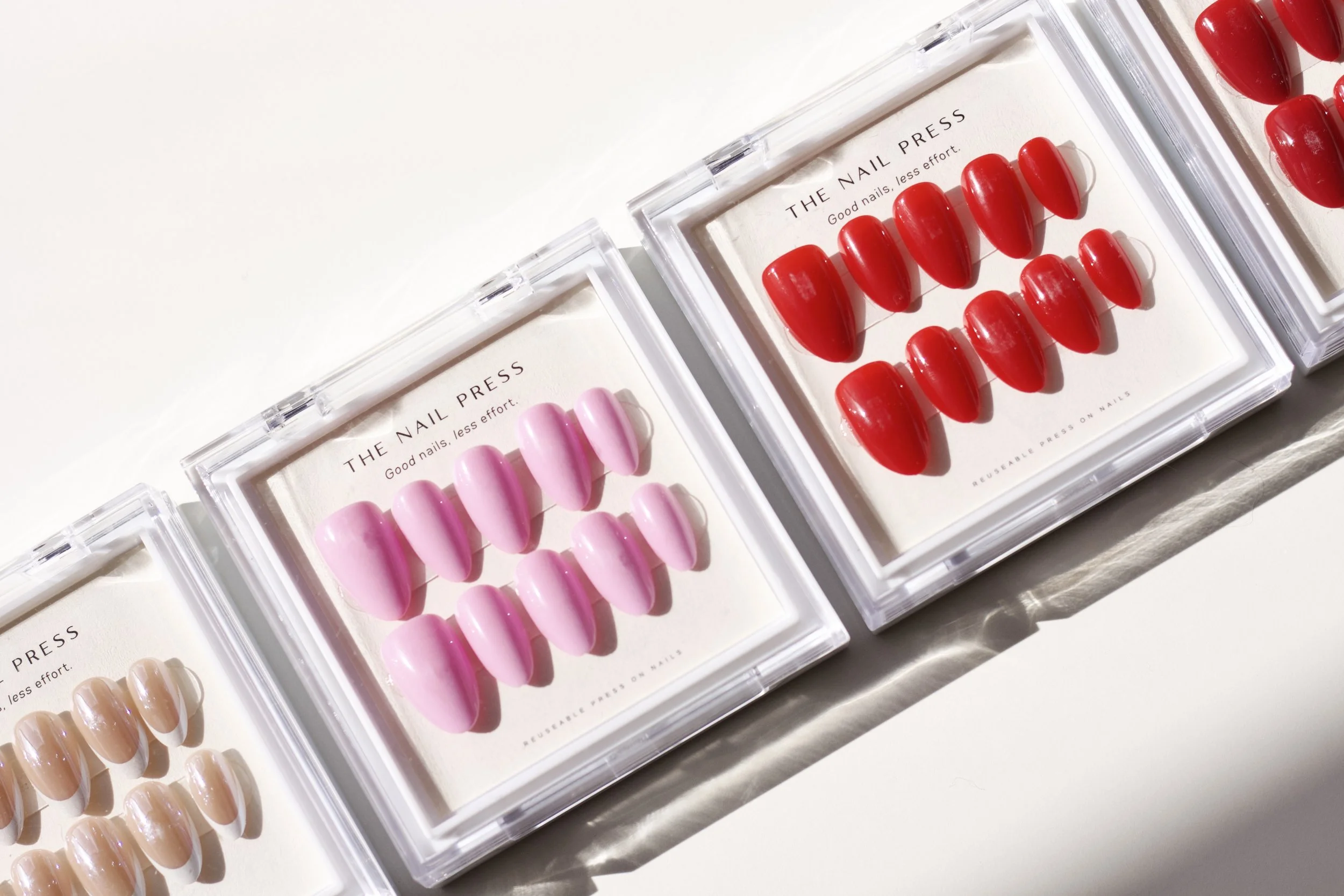 Display of three sets of colorful press-on nails in pink, red, and beige packaged in clear plastic cases with labels.