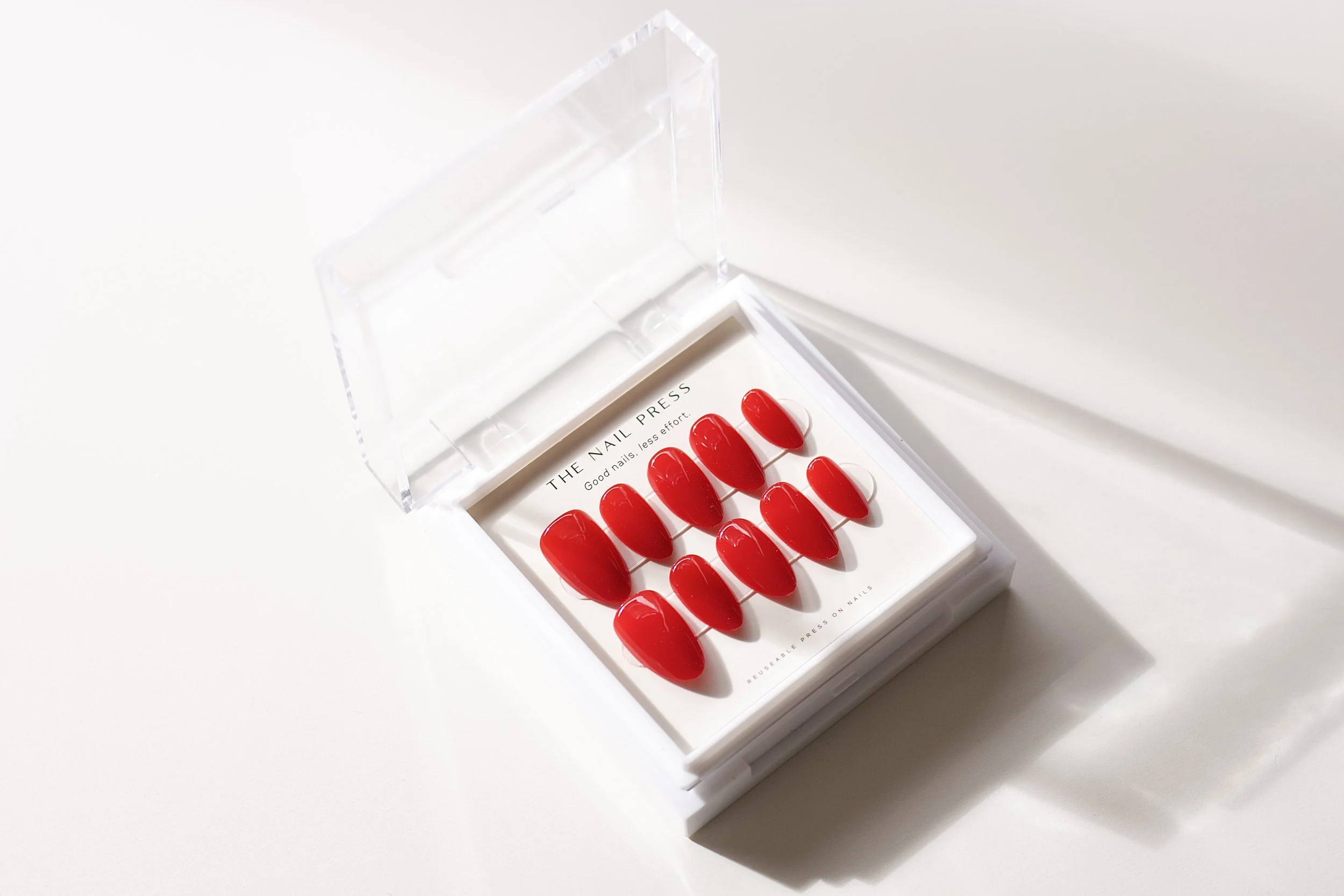 A clear plastic box containing red press-on nail tips arranged in two rows, with the text 'THE NAIL PRESS' and 'Good nails, less effort.' visible inside the box.
