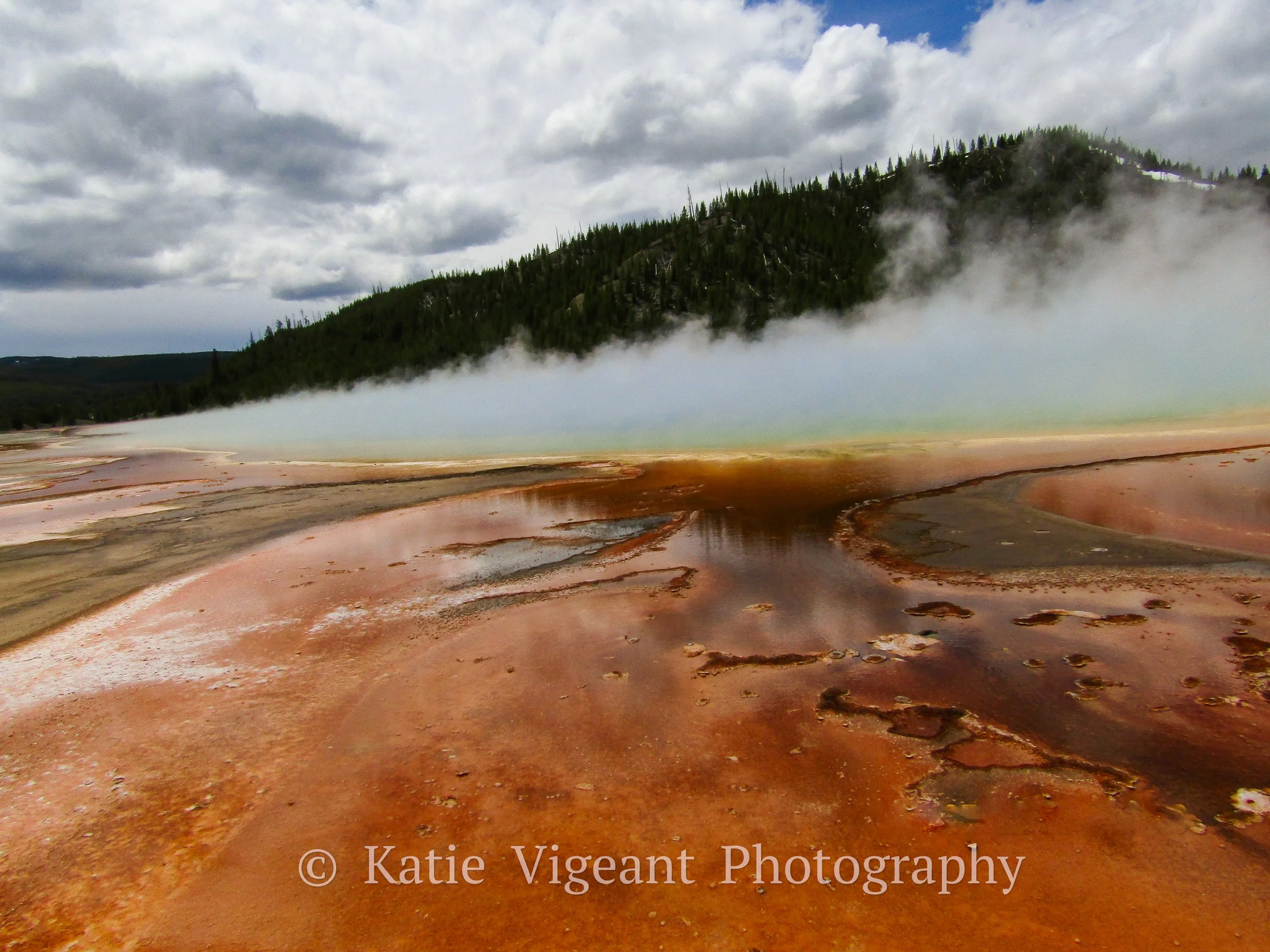 Colorful geothermal area with steam rising from hot springs, orange and pink mineral deposits, and a forested hillside in the background under cloudy skies.