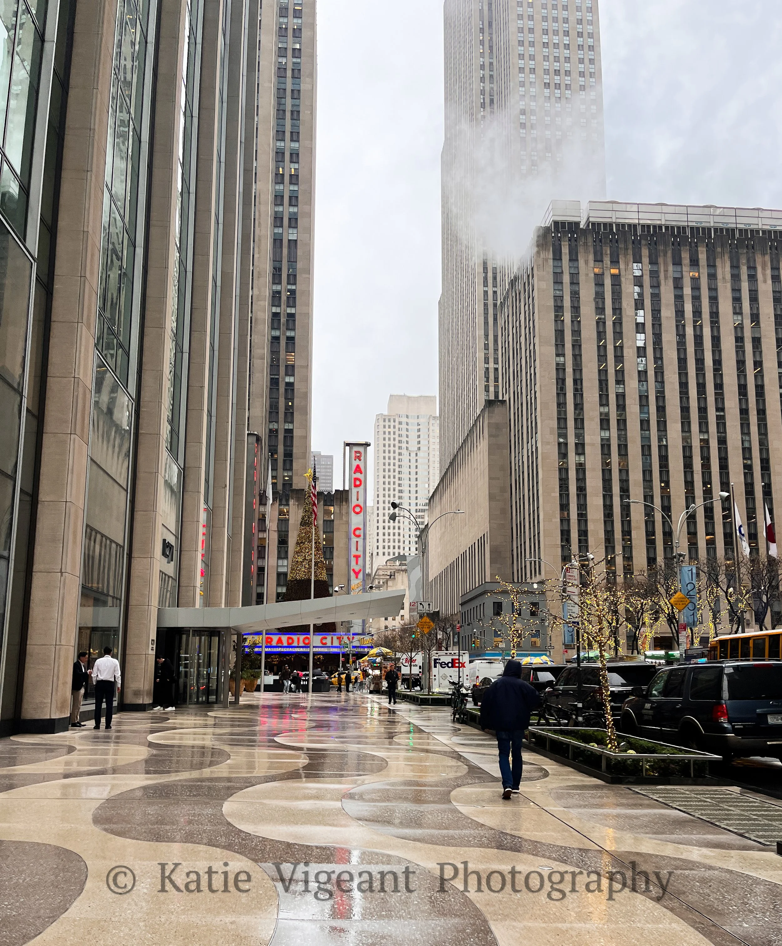 Downtown city street with tall buildings, a Radio City sign, people walking under wet weather, holiday decorations, and parked cars.
