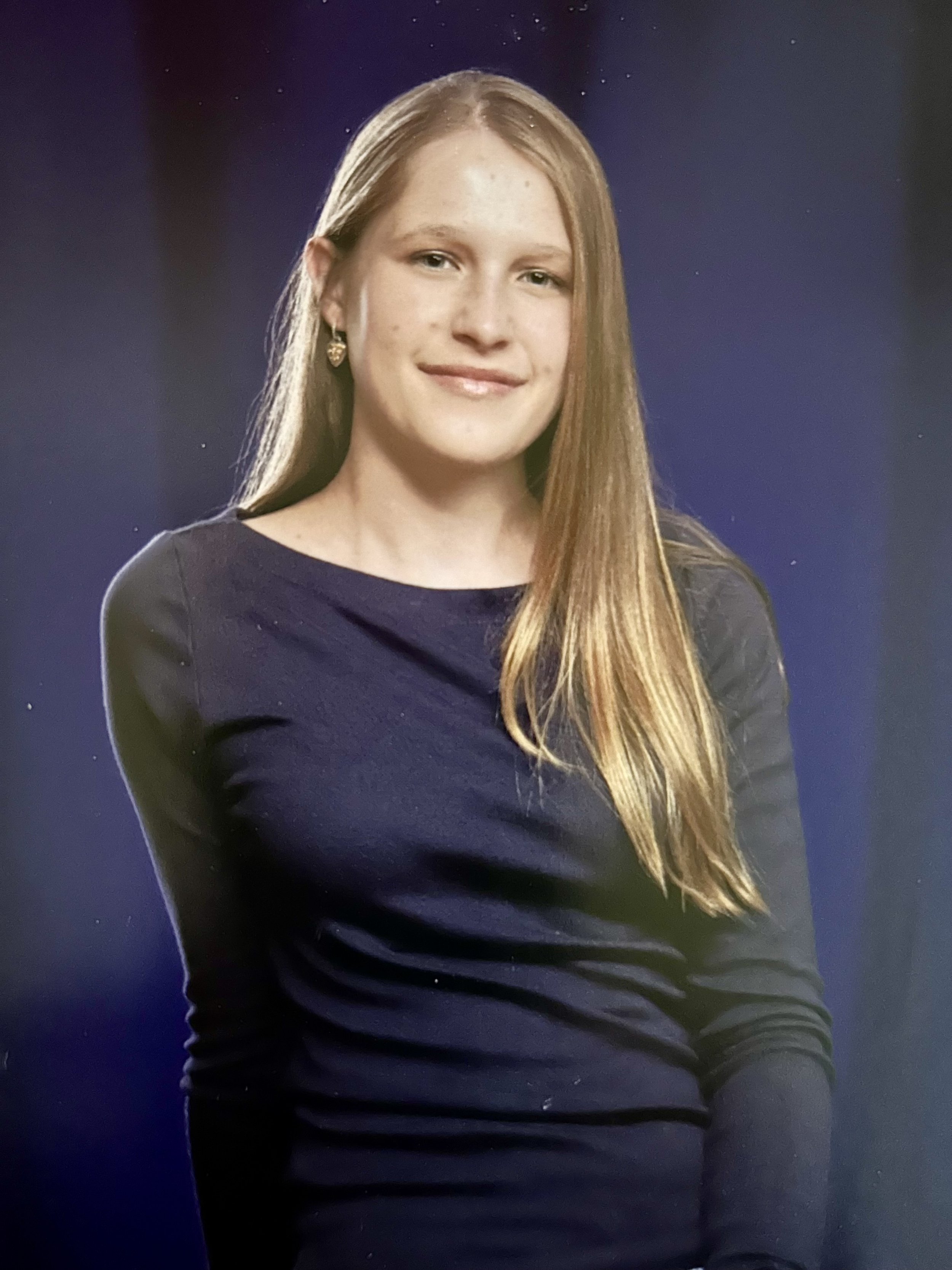 A young woman with medium-length blonde hair, wearing a dark long-sleeved shirt, smiling with earrings, against a dark background.