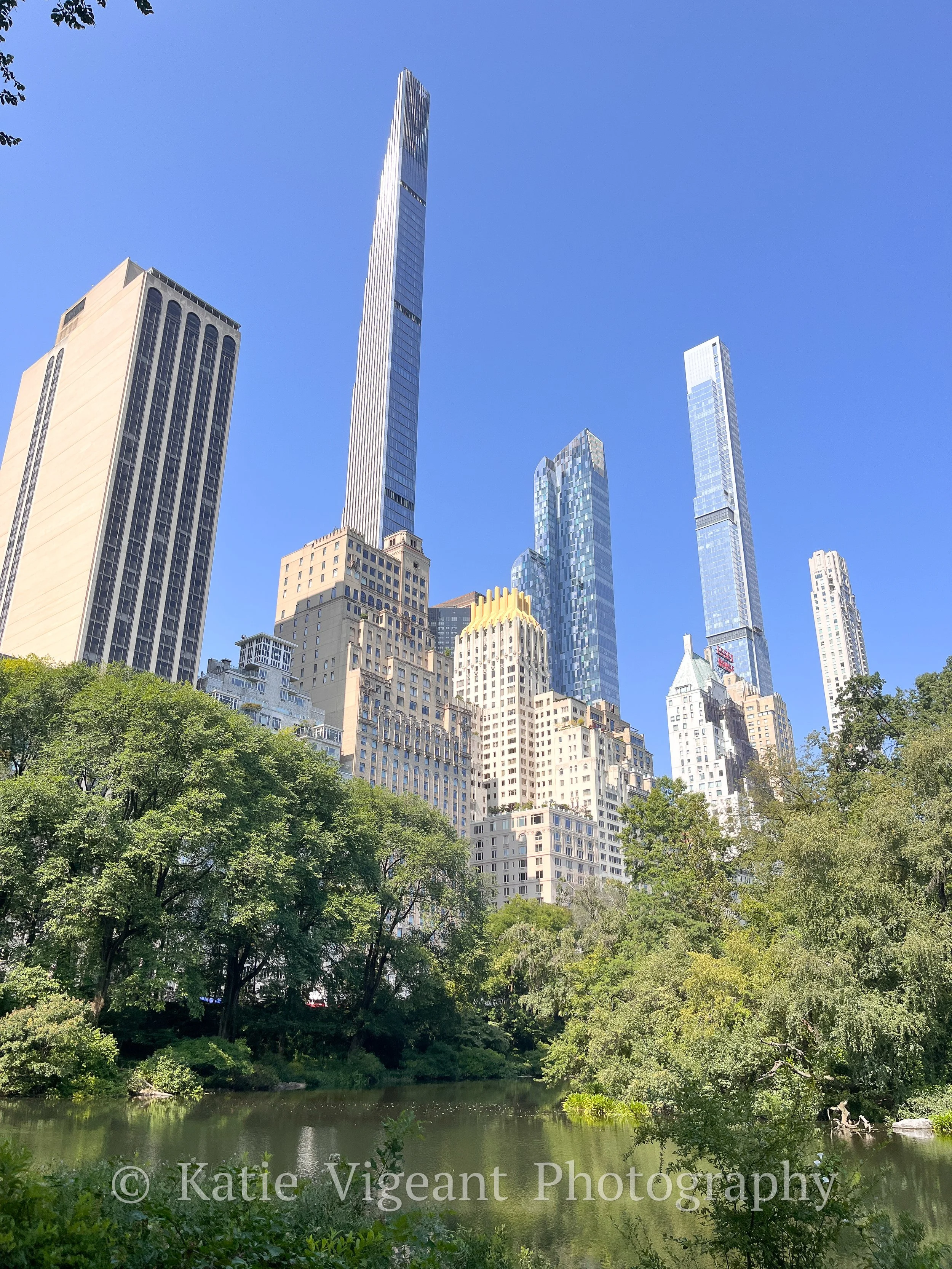 Skyscrapers in New York City viewed from Central Park with trees and a pond in the foreground and a clear blue sky.