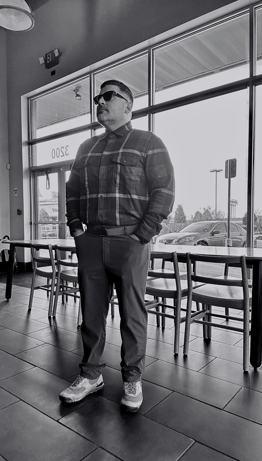 A man wearing sunglasses, a plaid shirt, and sneakers standing inside a cafe near large windows with parked cars outside.