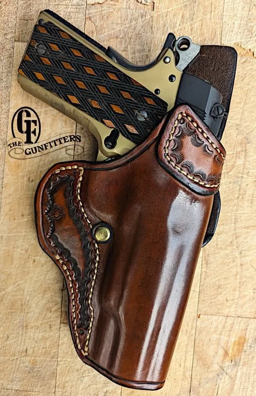 A semi-automatic pistol with a black and tan patterned grip in a brown leather holster, resting on a wooden surface with the logo 'The Gunfitters'.