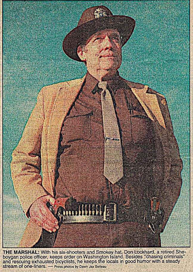 "The Marshall of Washington Island", WI, wearing a sheriff's badge, Smokey Bear style hat, a belt with a twin six-shooter holster, standing against a blue background.