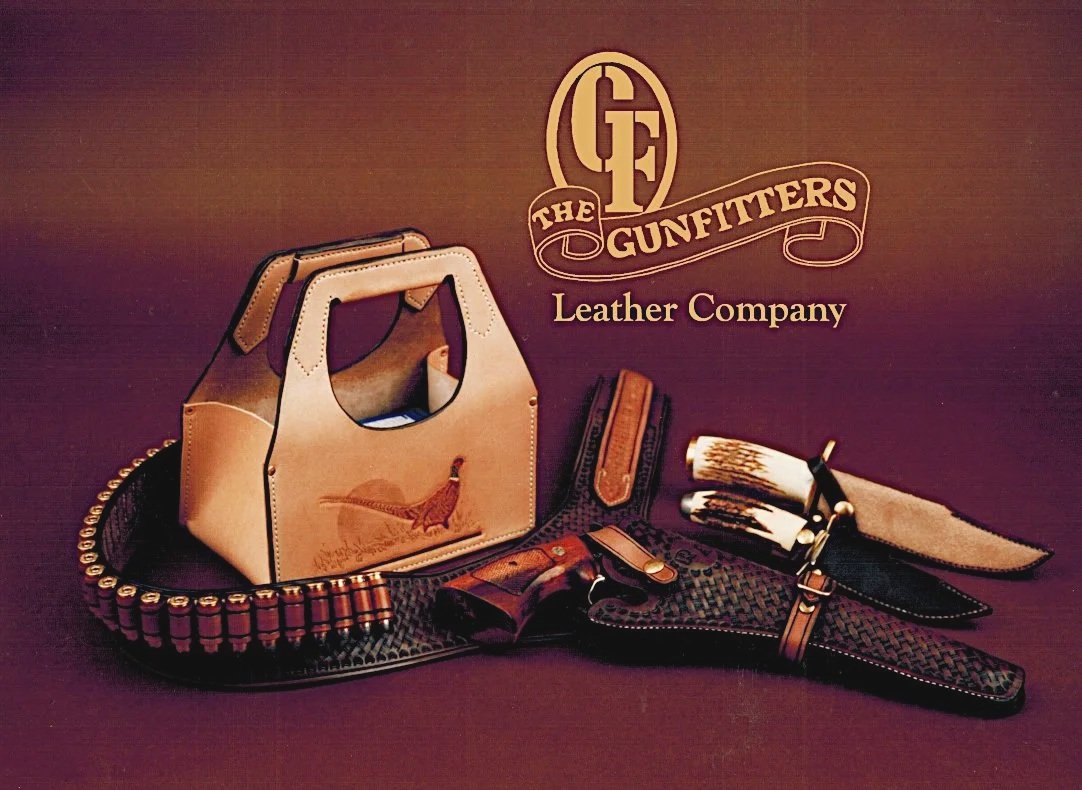 Display of leather accessories including a handbag, gun holster, belts, and a knife with a shearling sheath, with a logo reading 'The Gunfitters Leather Company' in the background.