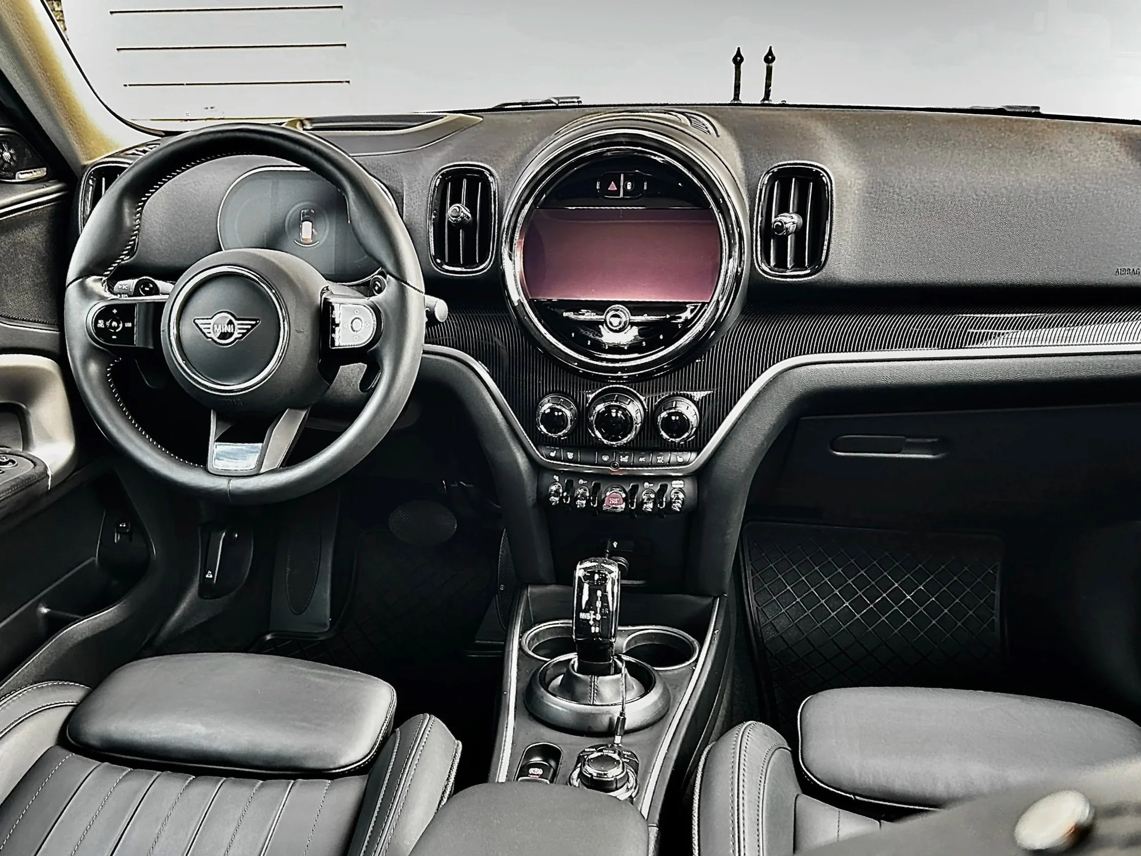 The interior of a Mini Cooper with a black dashboard, leather seats, and a central touchscreen display, with a gear shifter and other controls in the center console.