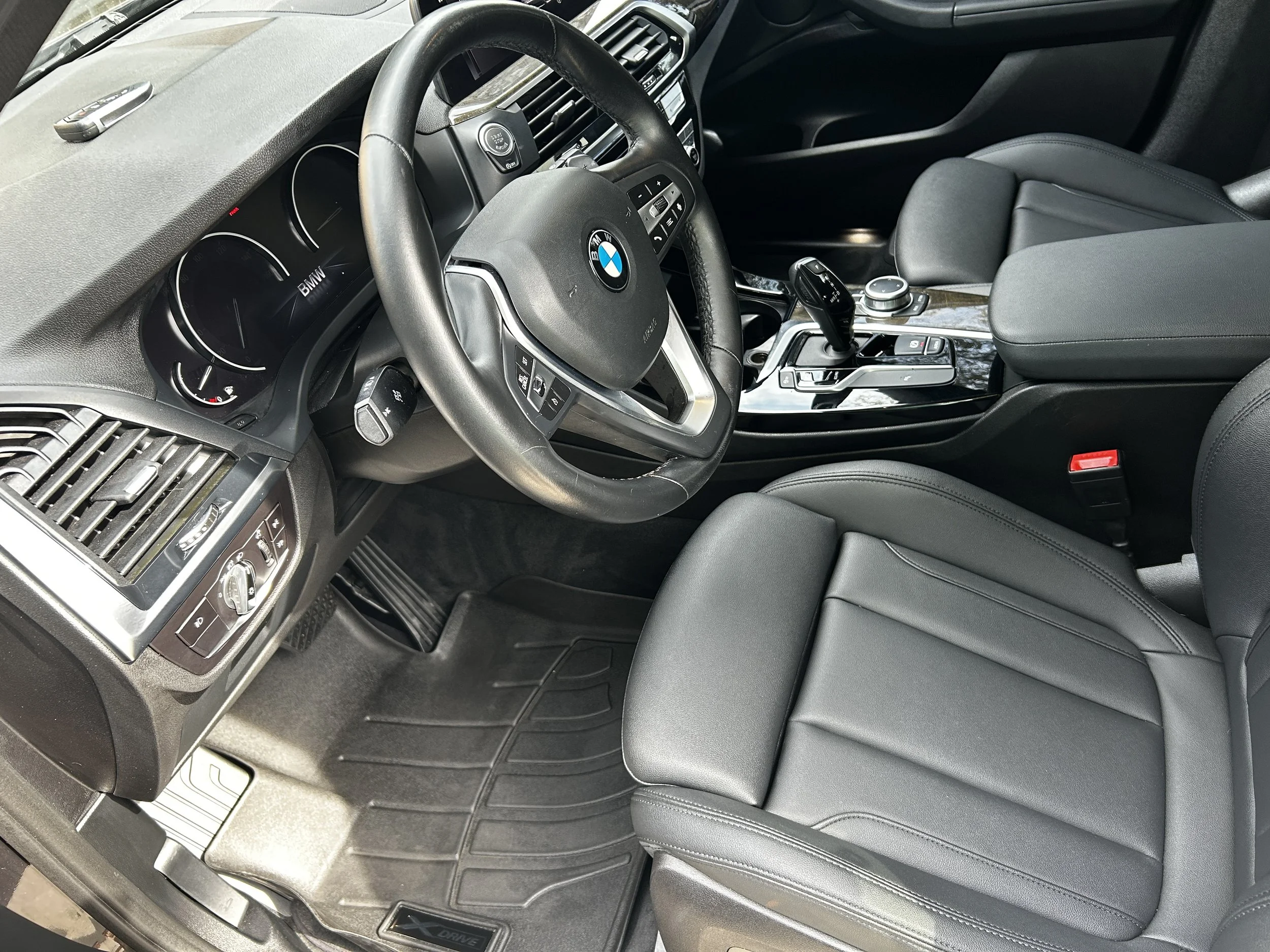 The interior of a BMW vehicle showing the steering wheel, dashboard, center console with gear shift, and black leather seats.
