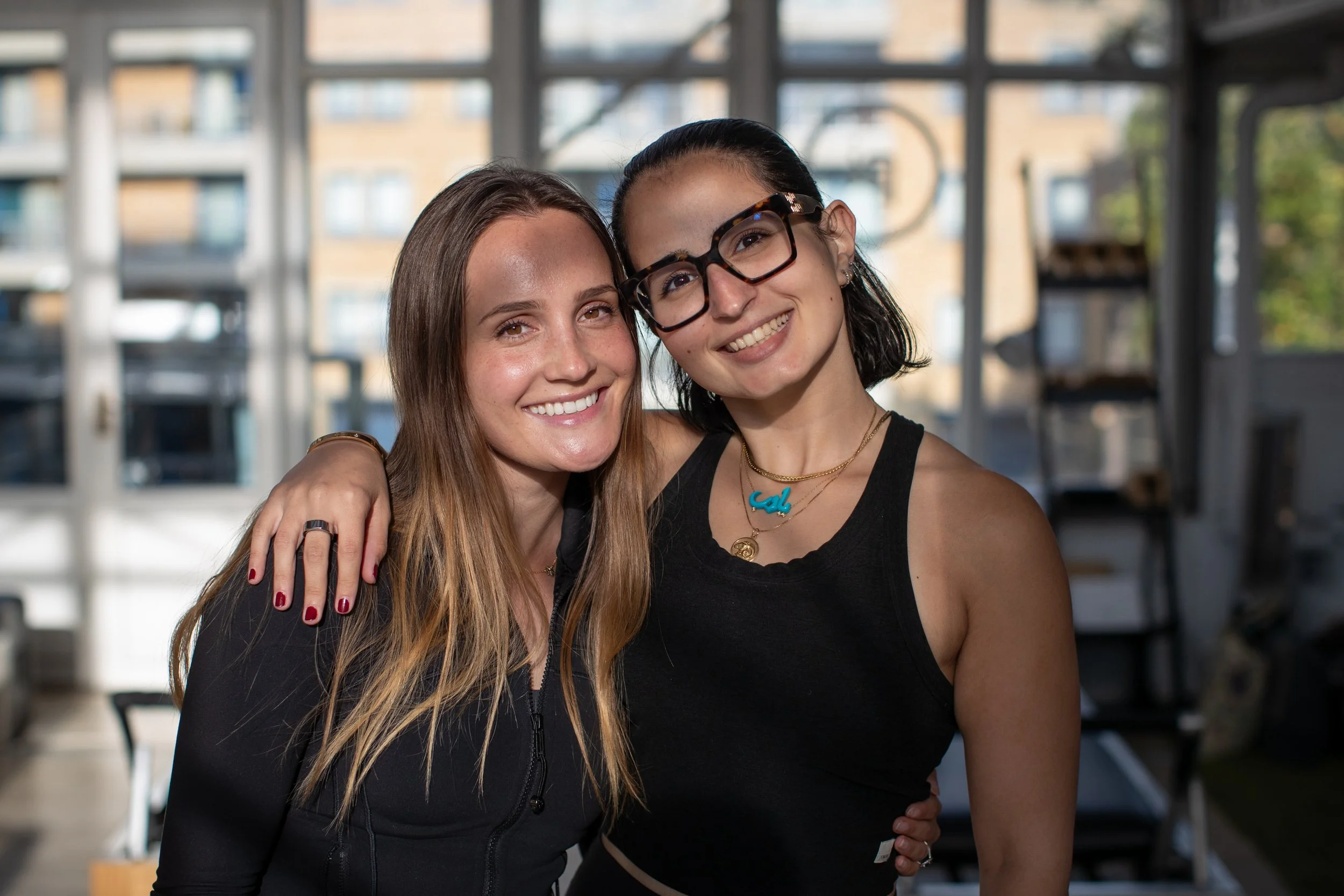 Two women smiling together indoors, embracing, with large windows and a cityscape in the background.