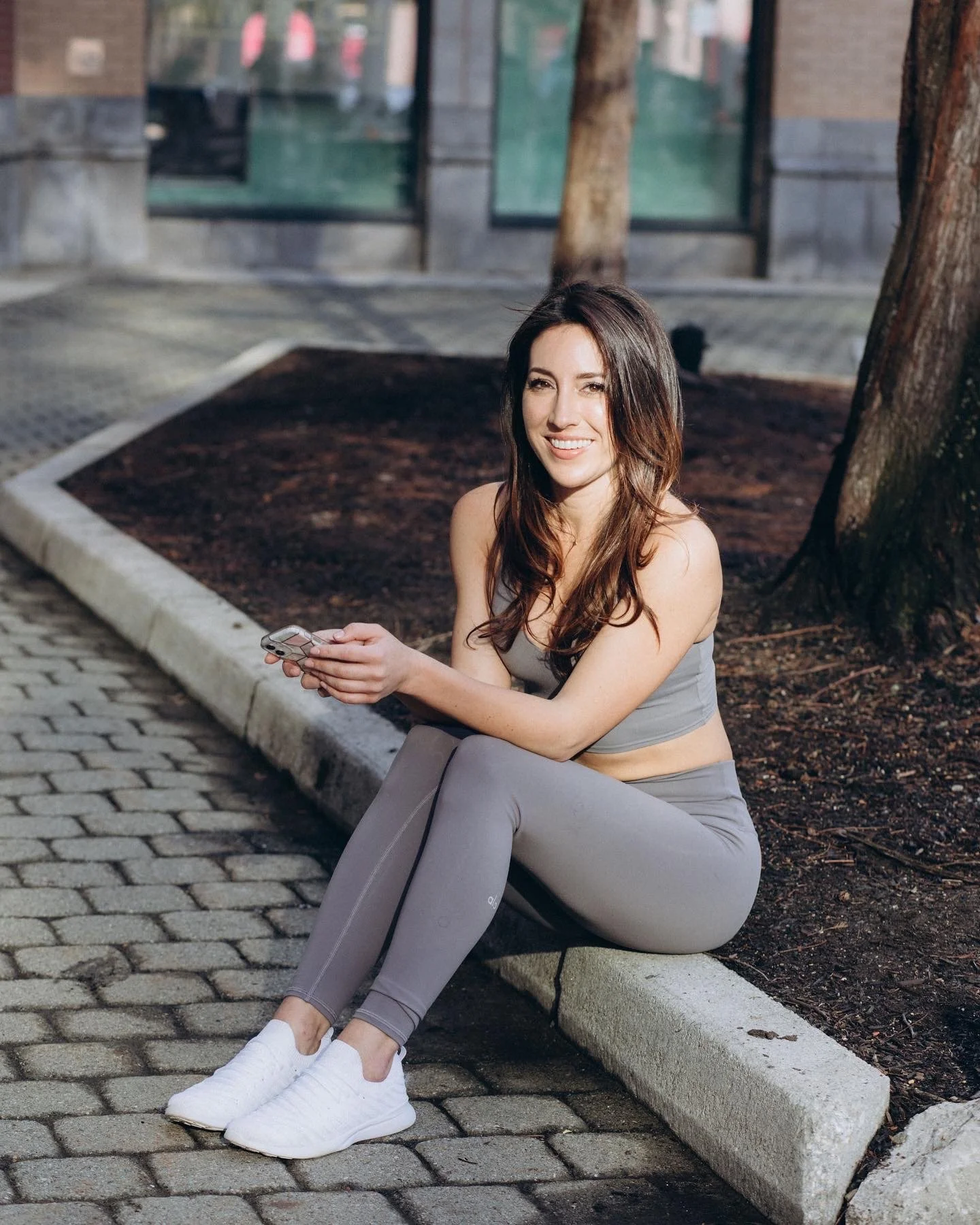 Young woman in workout clothes sitting on a curb in a park, holding a phone and smiling at the camera.