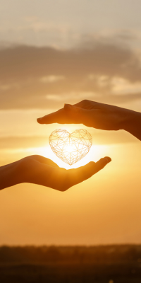Two hands holding a wireframe heart against a sunset background.