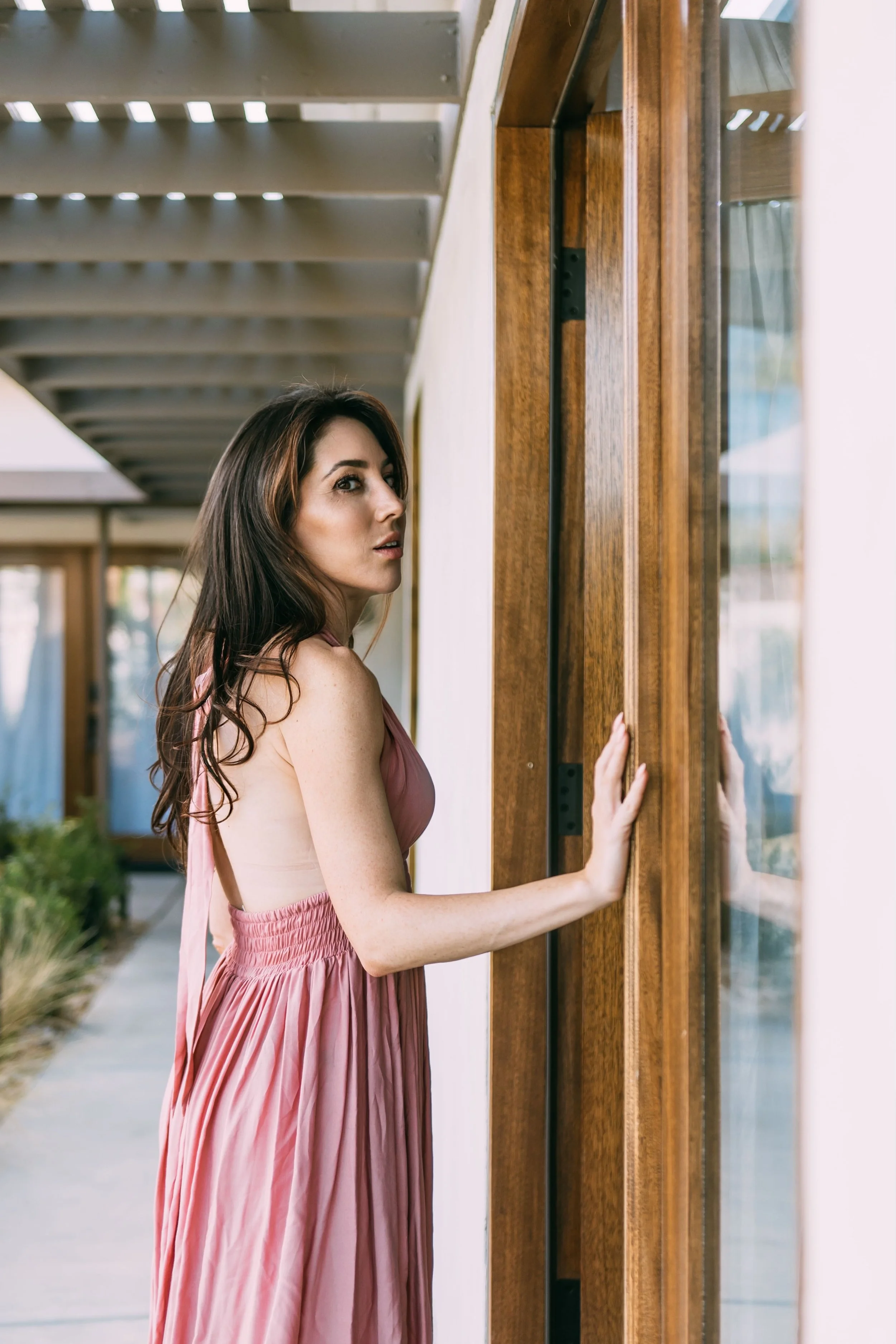 A woman with long dark hair wearing a pink dress looking at a glass door outside.
