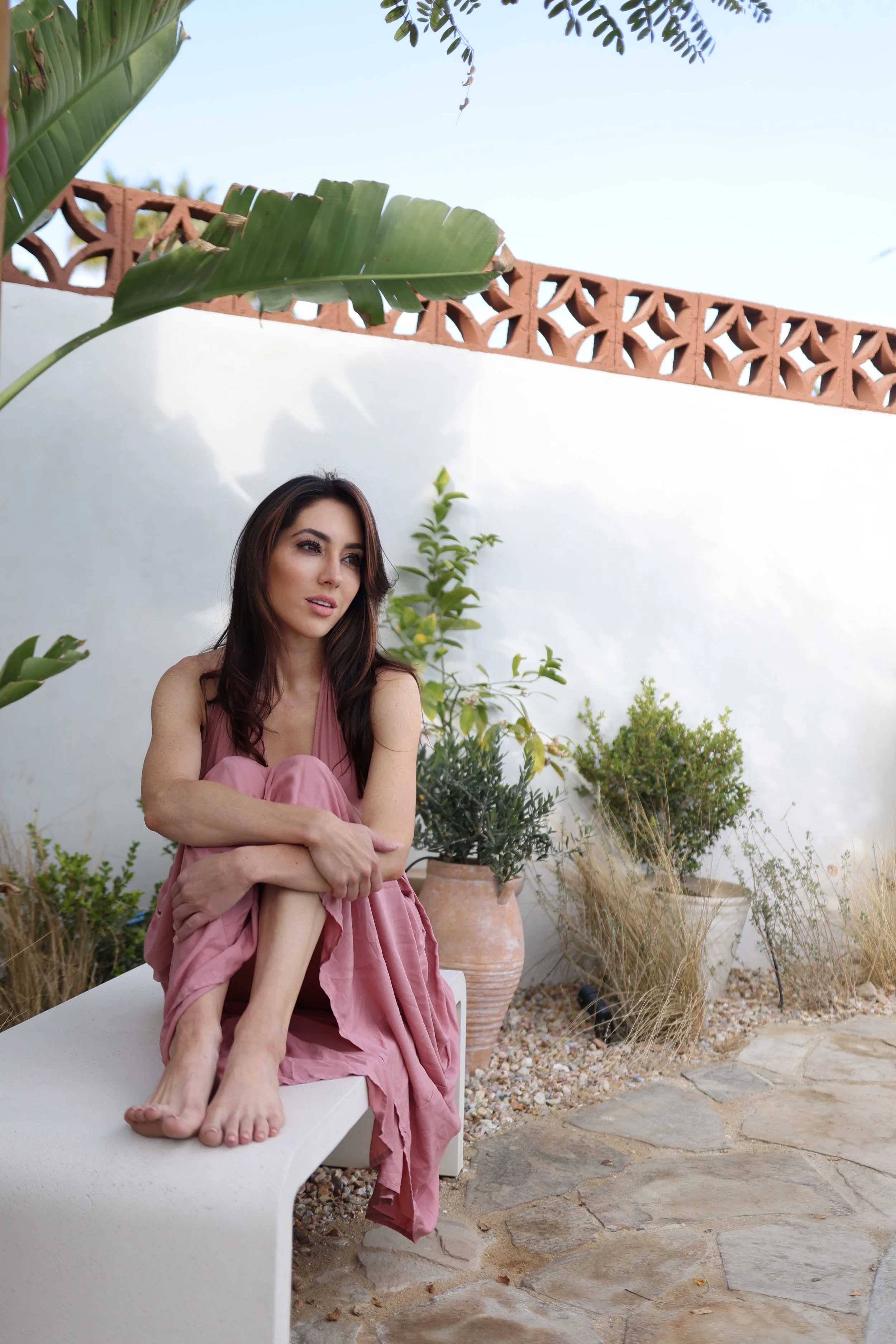 A woman sitting on a white outdoor bench in a garden with potted plants, a white wall, and a decorative brick wall in the background, wearing a pink sleeveless dress.