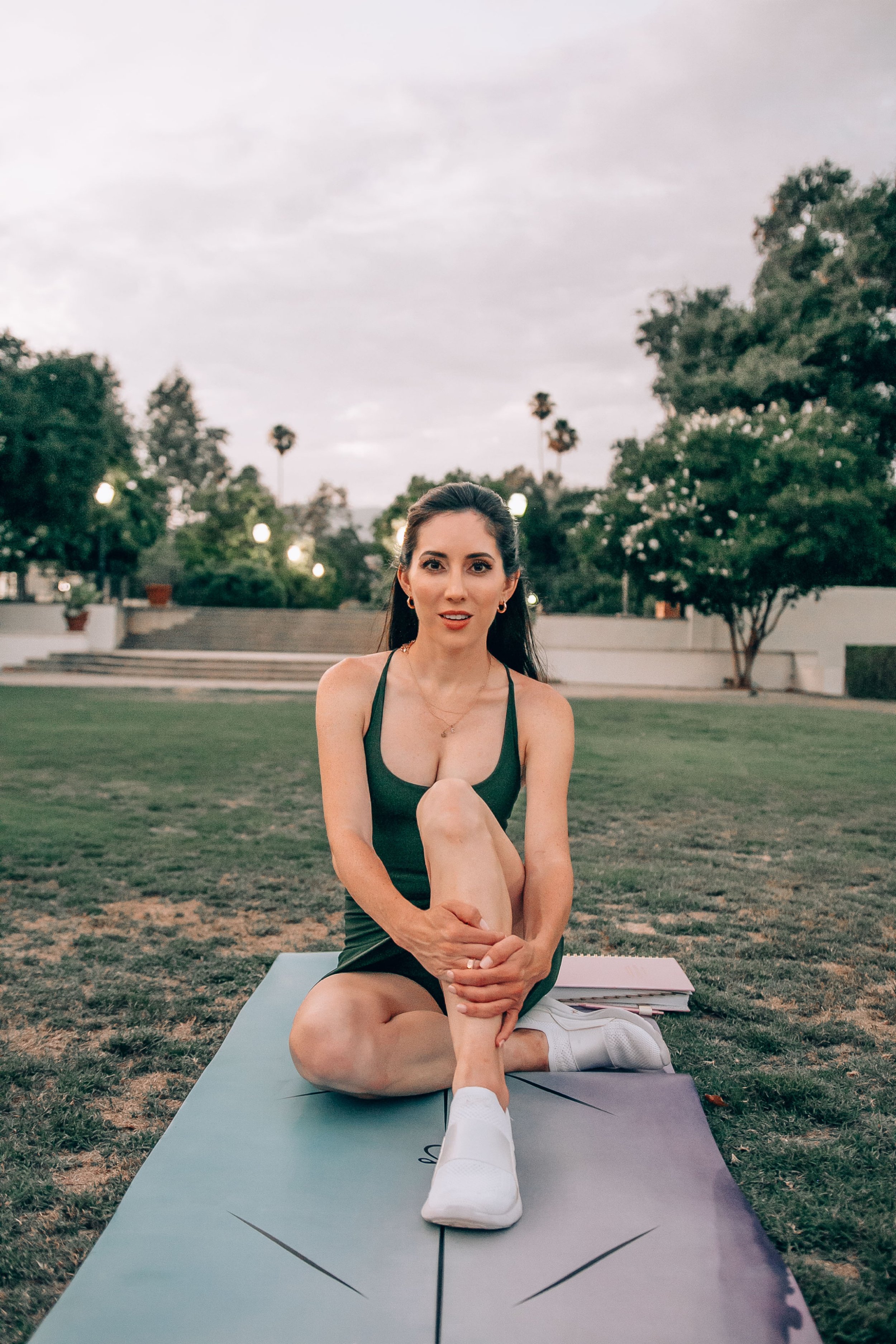 A woman in athletic clothes practicing yoga on a mat outdoors in a park during the evening.