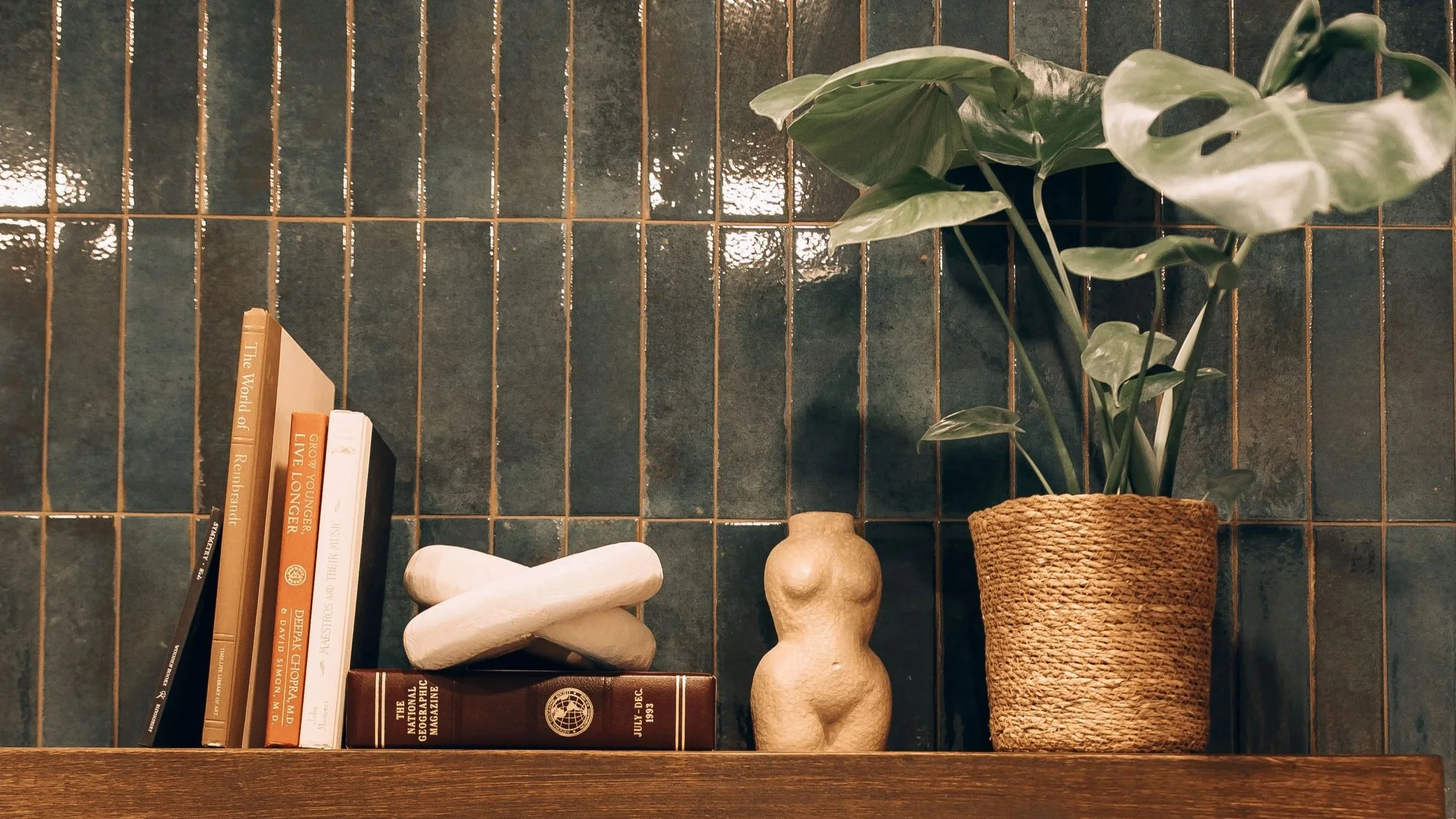 Shelf with books, a small abstract sculpture, a ceramic torso, and a potted plant against a dark tiled wall.
