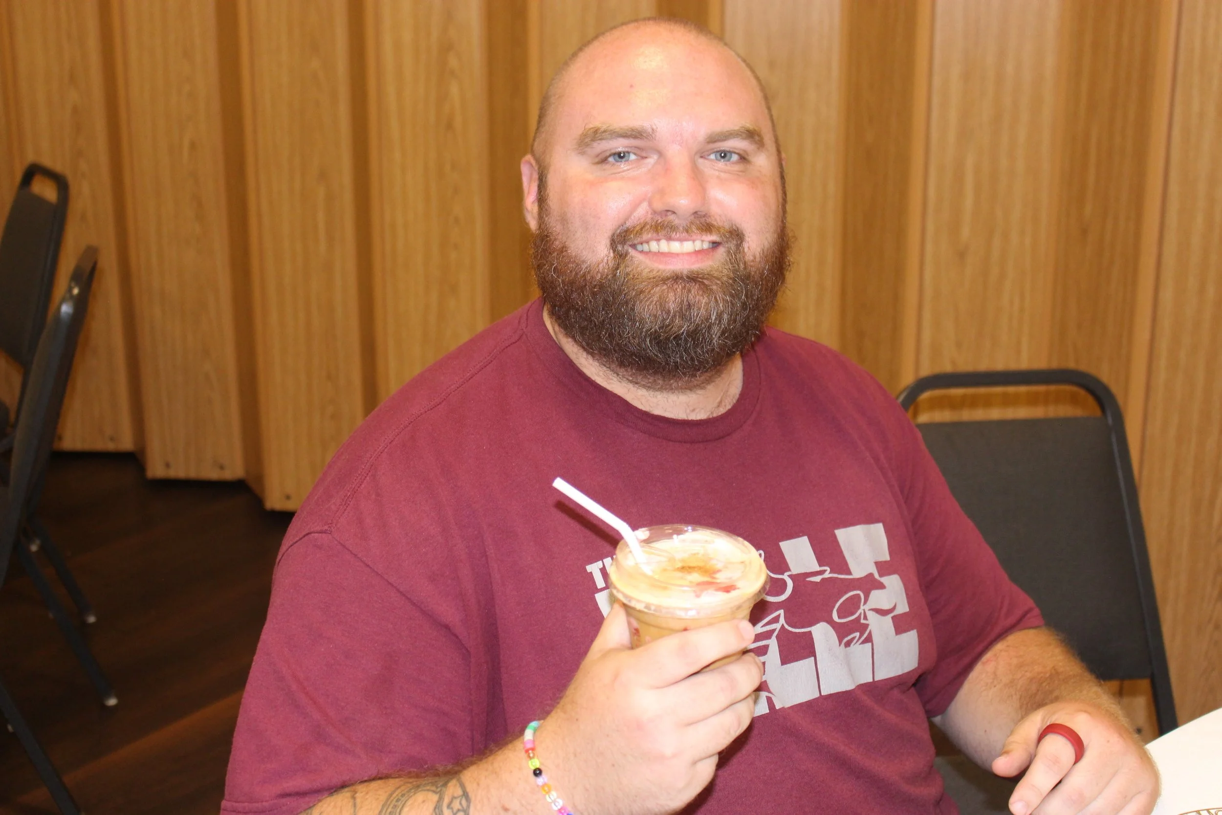 A man with a beard and light blue eyes holding a dessert with a straw, smiling at the camera, wearing a maroon shirt in a room with wooden wall paneling and black chairs.