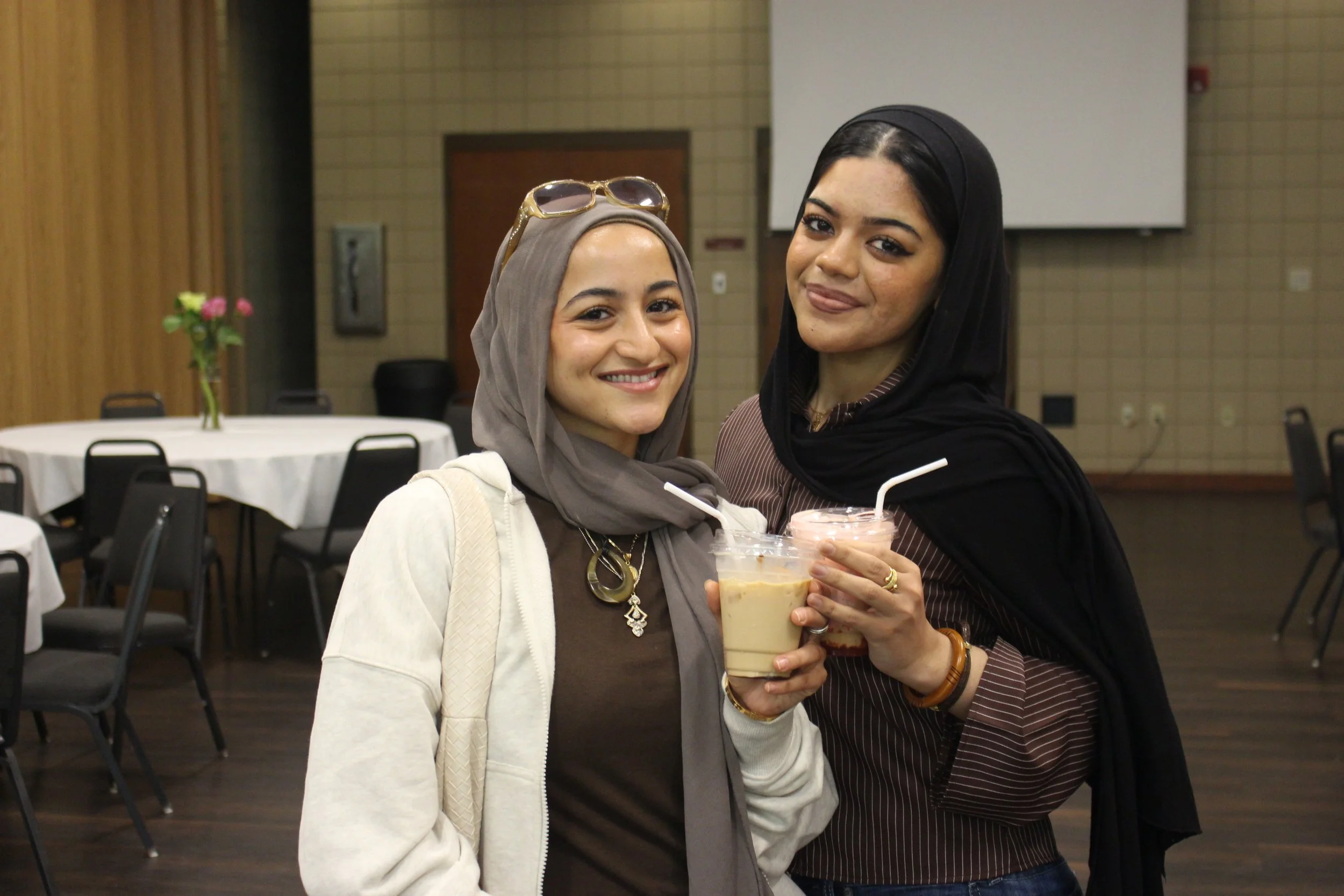 Two women smiling and holding drinks in a banquet hall with tables and chairs in the background.