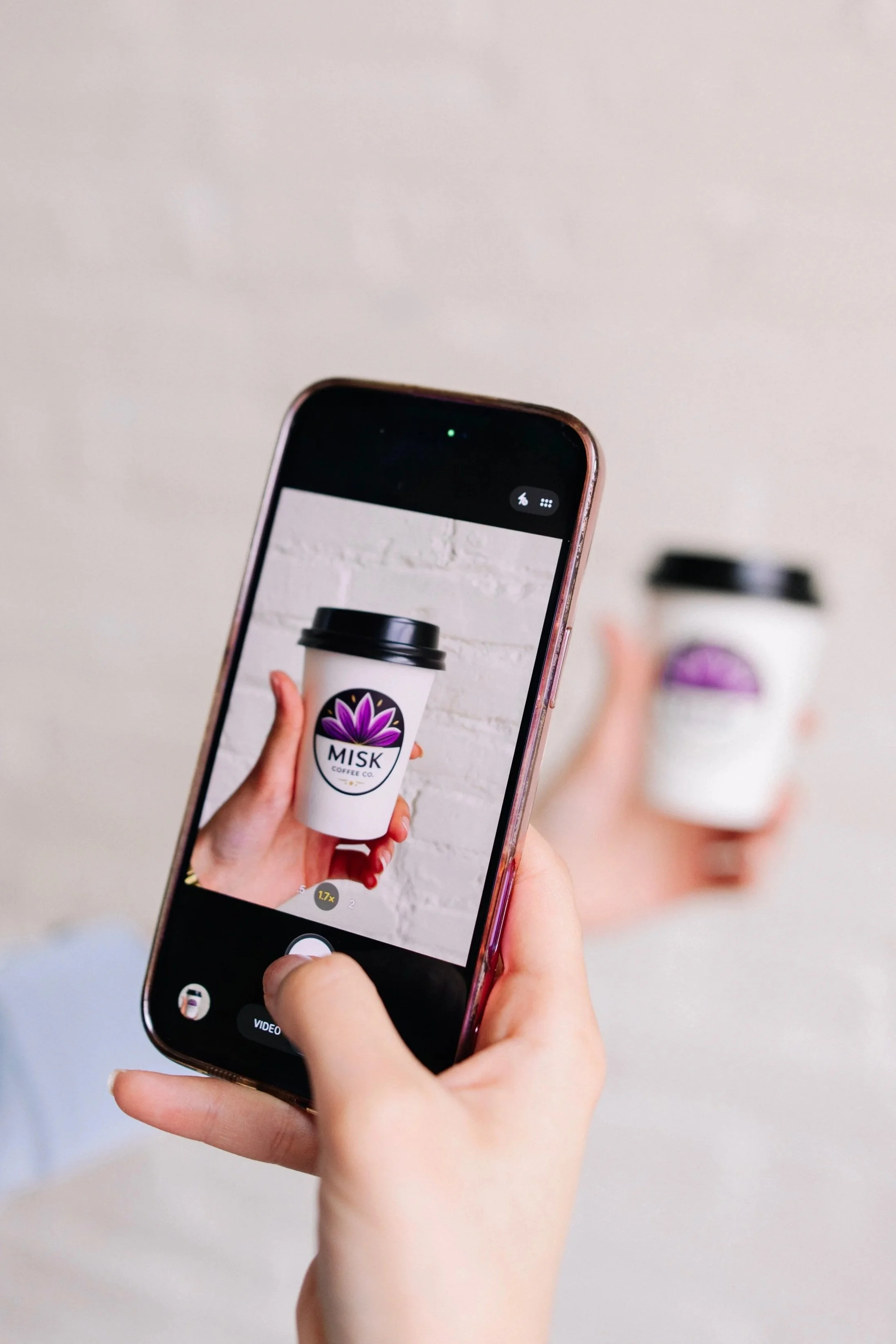 A person is taking a photo of a disposable coffee cup with a purple lotus flower logo and the text IASK covered with a black lid, using a smartphone with a rose gold case. The background is blurred, showing another similar coffee cup.