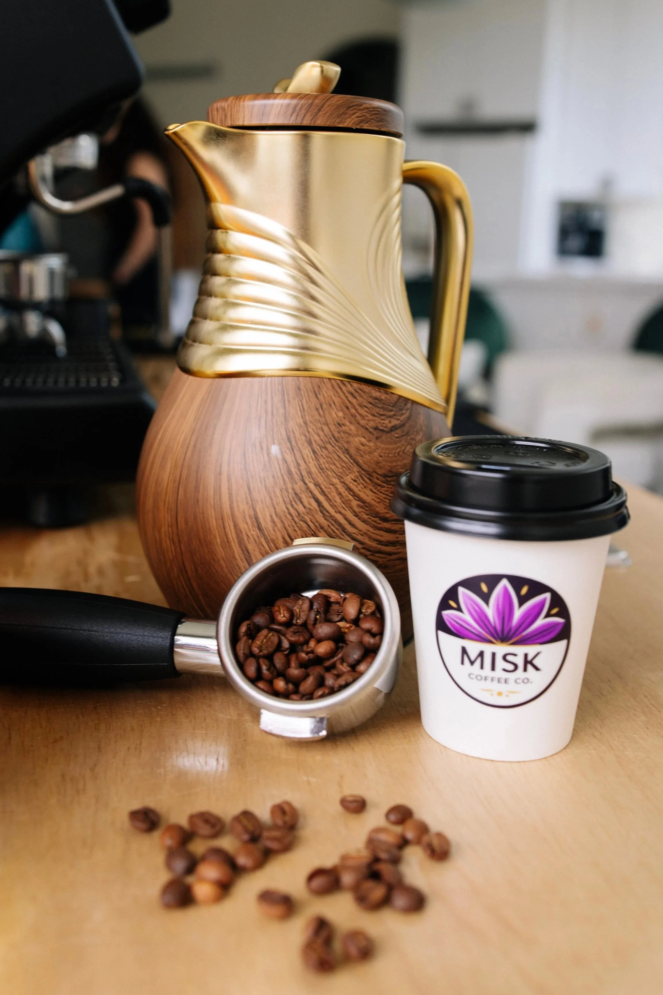 A wooden and gold-colored decorative coffee pot, a Starbucks coffee cup with a lid, a portafilter filled with coffee beans, and a small container of coffee beans on a wooden surface.