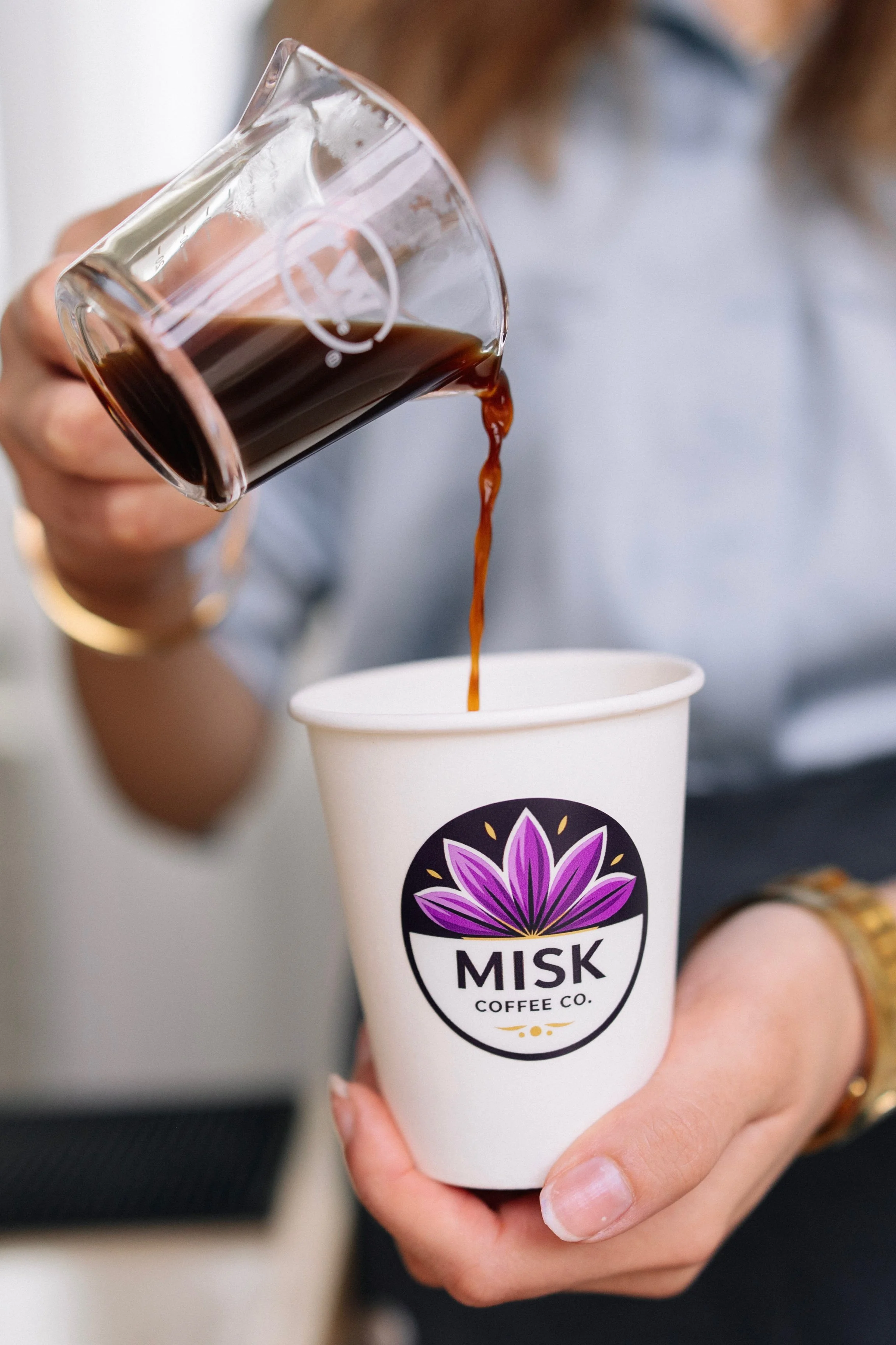 A person holding a white paper cup labeled 'MASK COFFEE CO.' with a purple flower logo, pouring black coffee into the cup.