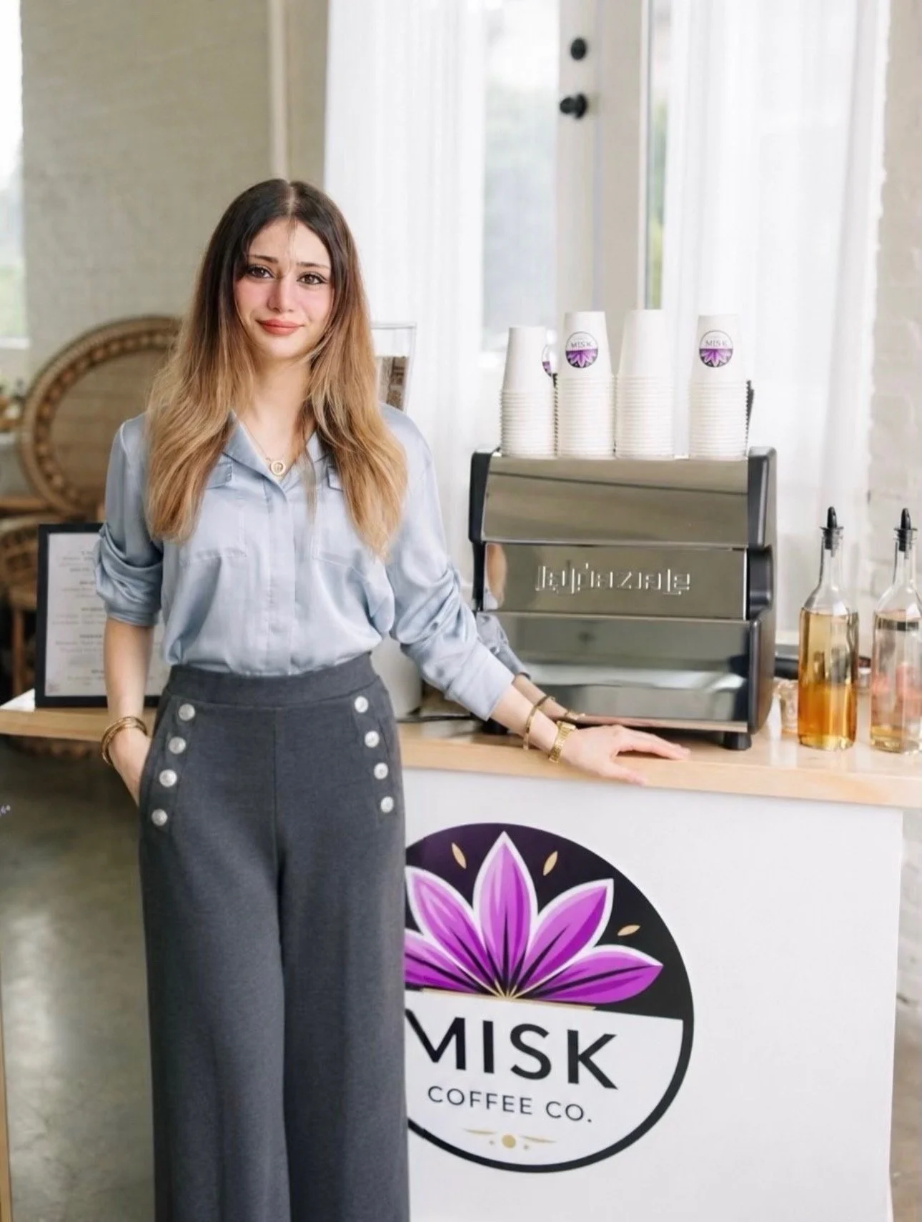 A woman standing behind a coffee bar at Mishka Coffee Co., wearing a light blue shirt and gray high-waisted pants with buttons, smiling and posing with her right hand resting on the counter.