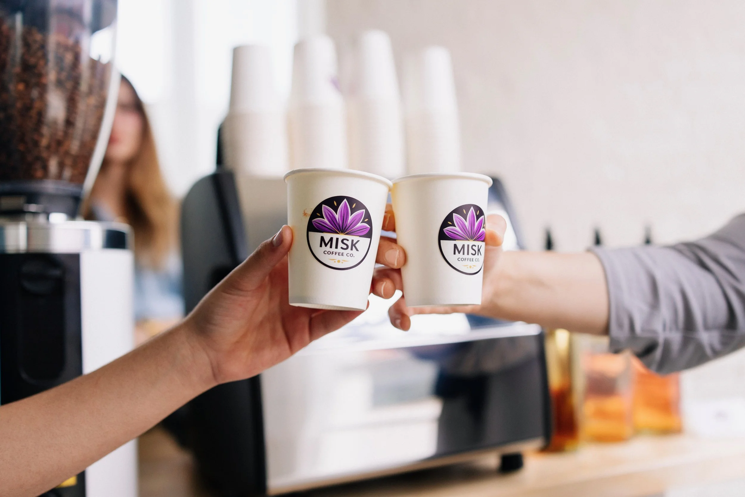 Two people clinking white coffee cups with a purple and black logo that reads 'MISK Coffee Co.' inside a coffee shop, with an espresso machine and stacks of paper cups visible in the background.