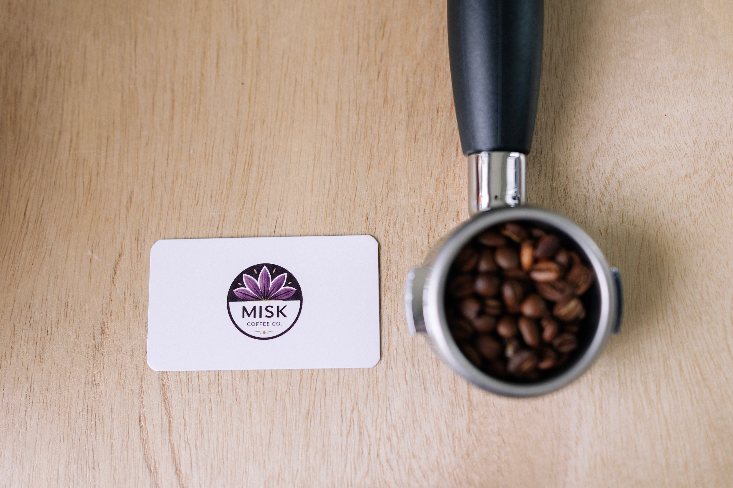 Close-up of a coffee grinder with coffee beans inside, placed on a wooden surface next to a white card with a purple flower logo and the text "MISK Coffee Co."