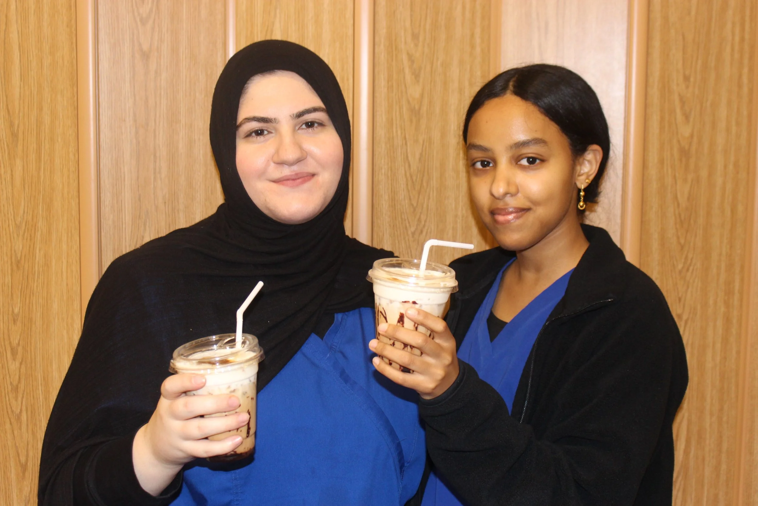 Two women holding milkshakes with straws, standing in front of a wooden wall. The woman on the left wears a black hijab and the woman on the right has short black hair and gold earrings.