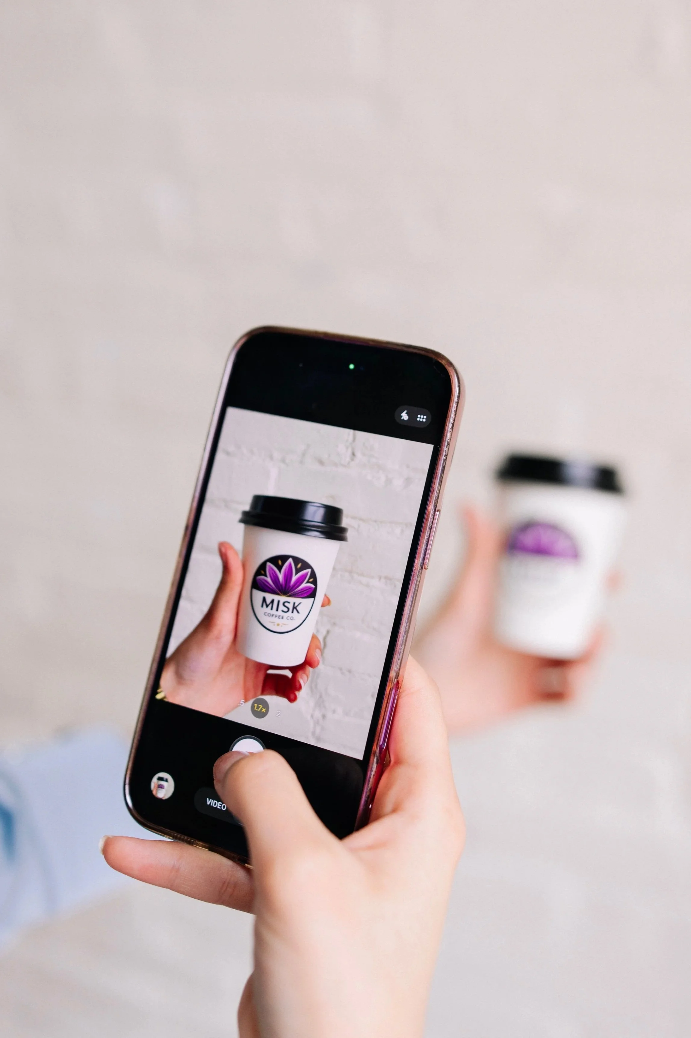 A person holding a smartphone displays a photo of a white coffee cup with a black lid and a purple flower logo on it, against a white brick wall background.